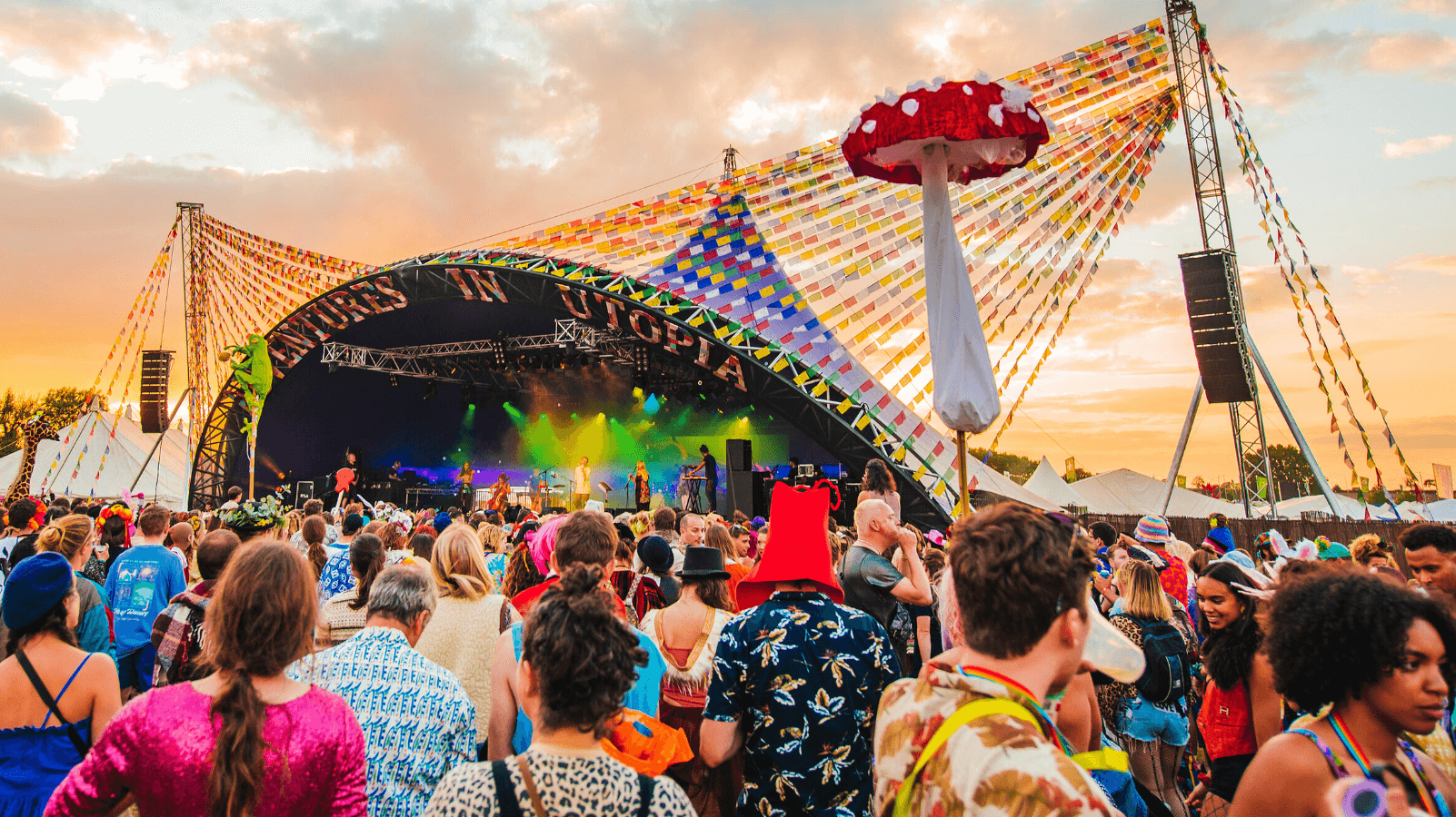 Crowd at a vibrant outdoor music festival with a colorful stage and decorative mushroom under a sunset sky.