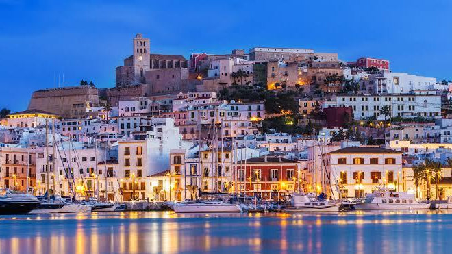 Dalt Vila's historic white buildings and cathedral illuminated at dusk above the Ibiza harbor with moored yachts.