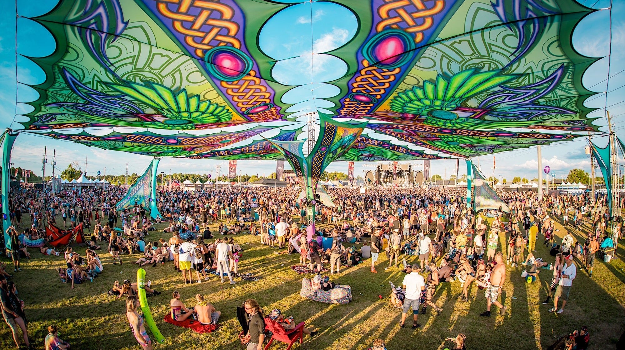 Vibrant festival crowd under colorful, patterned shade structures at an outdoor music event.