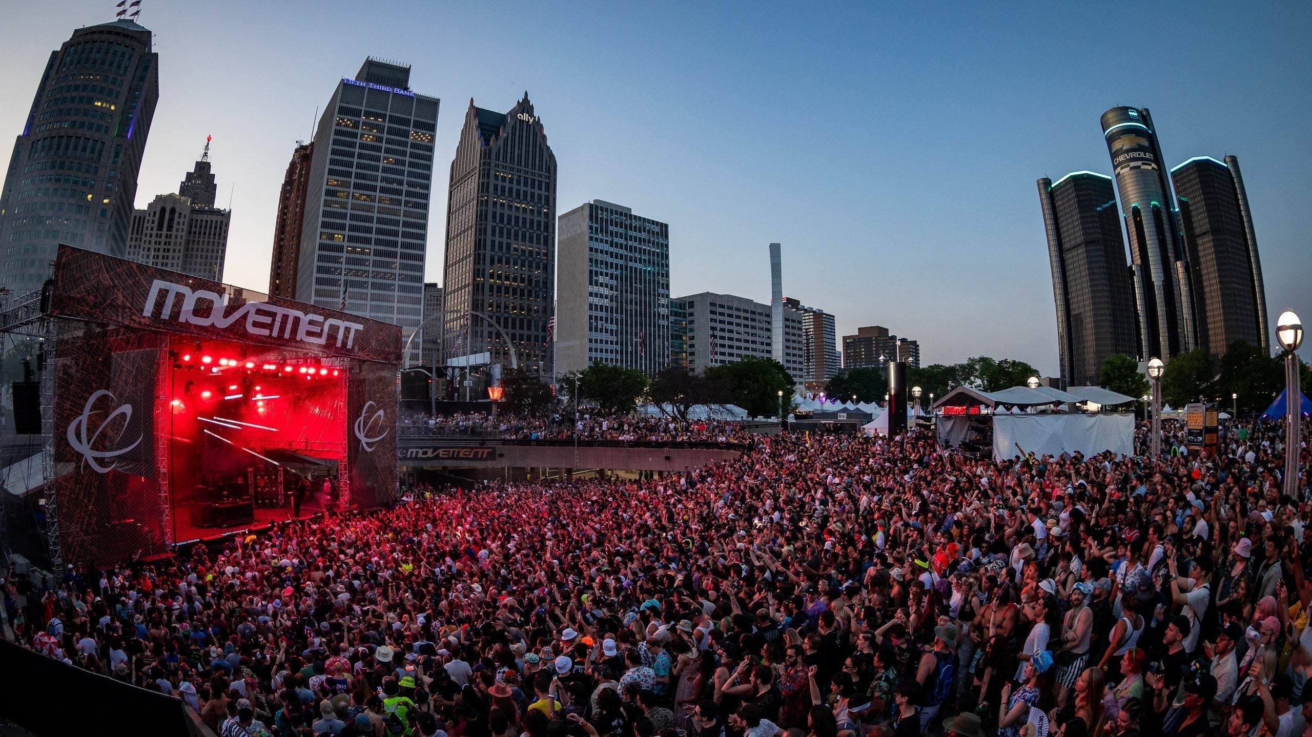 Crowd at Movement Detroit festival in Hart Plaza with city skyline at dusk, red stage lights.