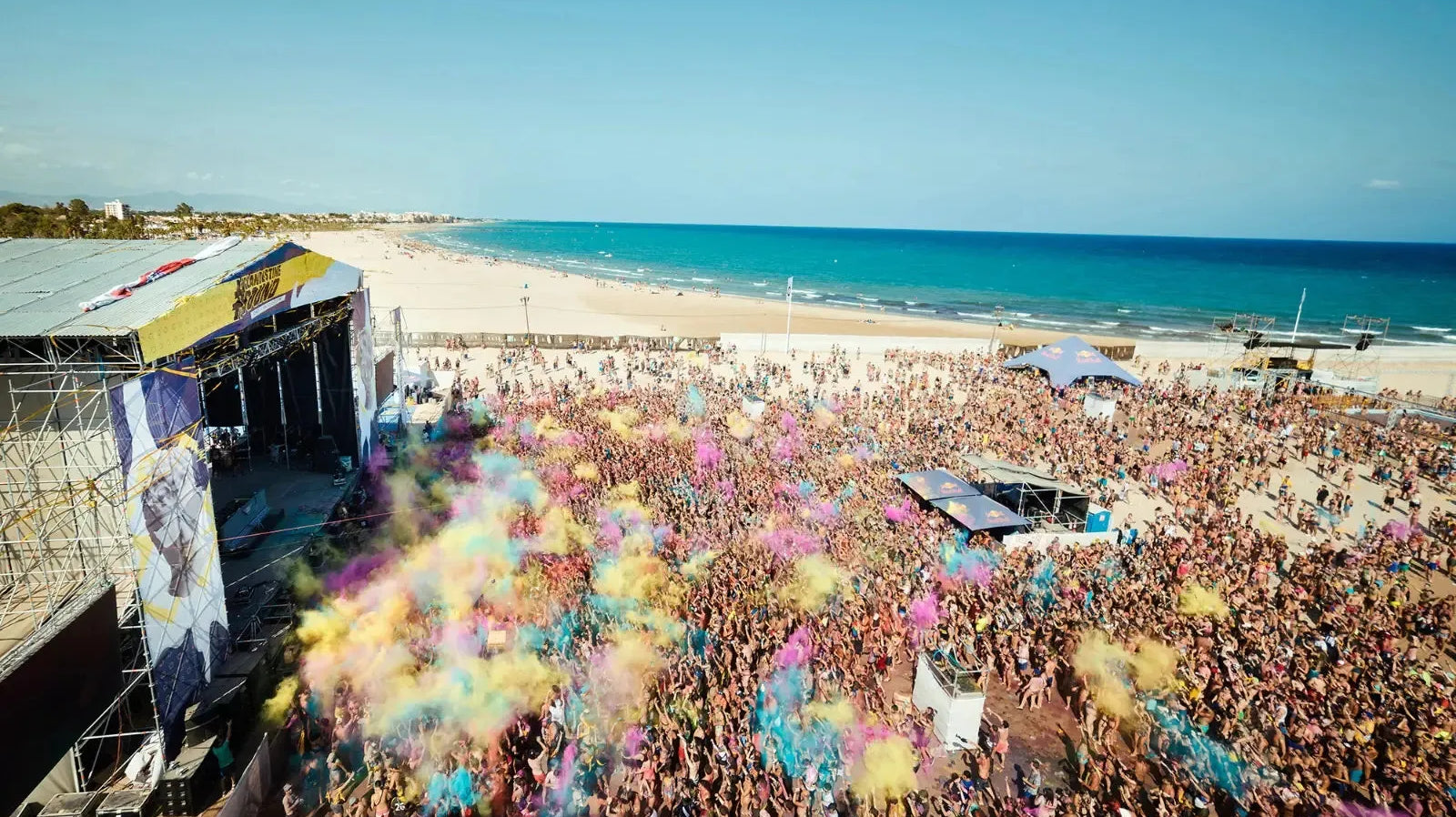 Arenal Sound beach festival with a large crowd throwing colorful powder, a stage, and the blue sea.
