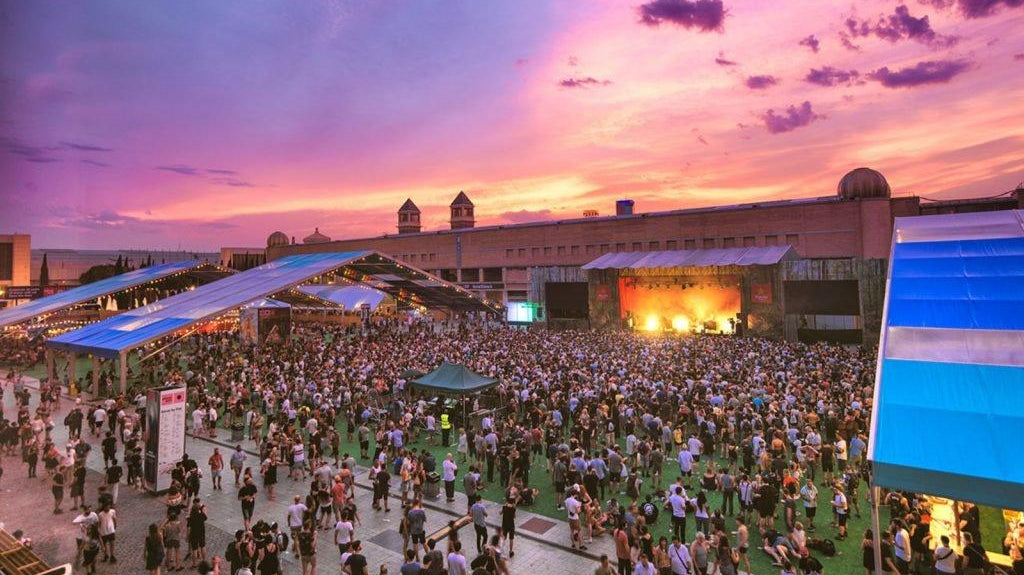 Large music festival crowd under a vibrant purple and orange sunset sky with a stage and tents.