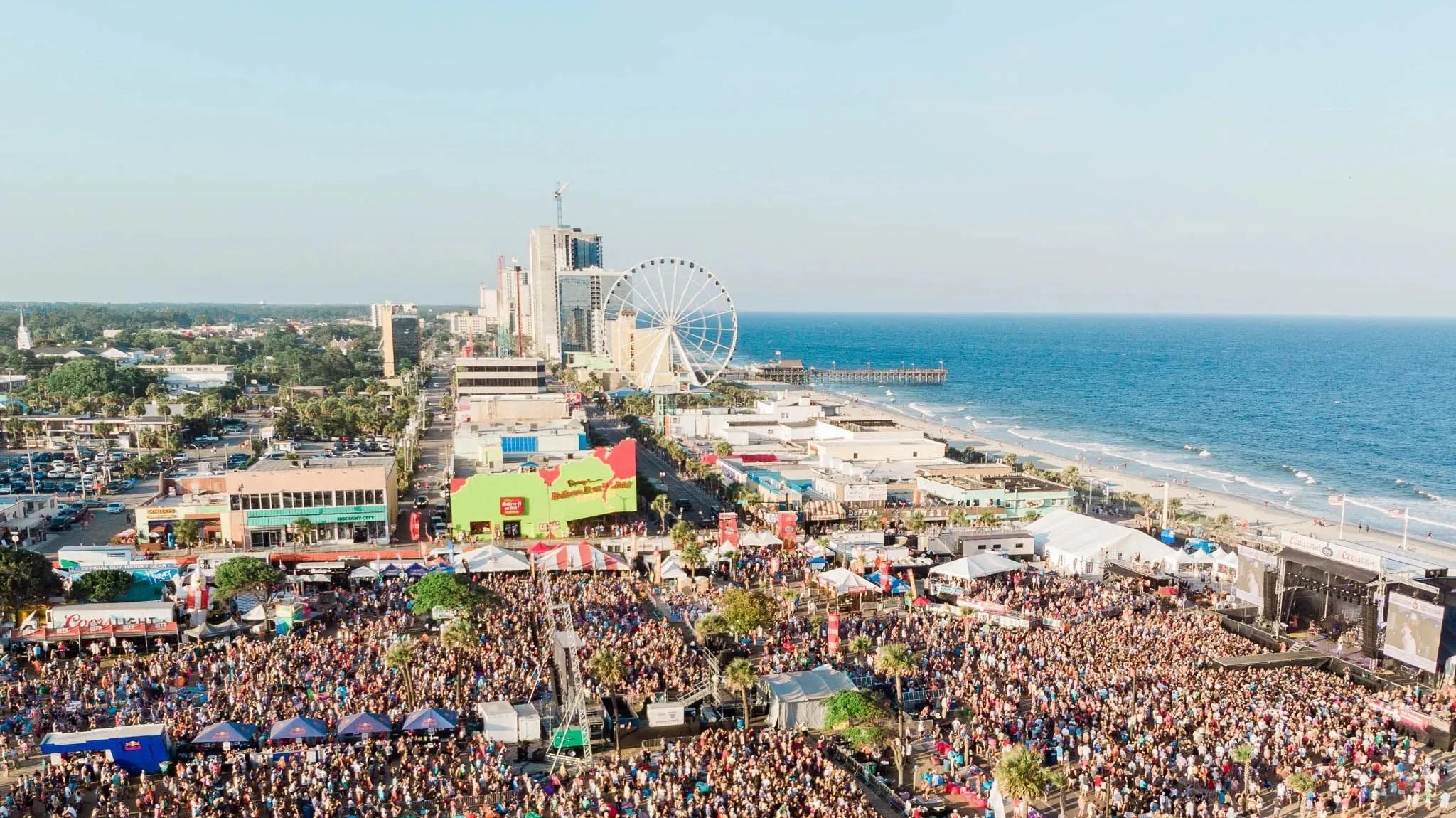Aerial view of a crowded outdoor music festival on Myrtle Beach with a Ferris wheel and ocean.