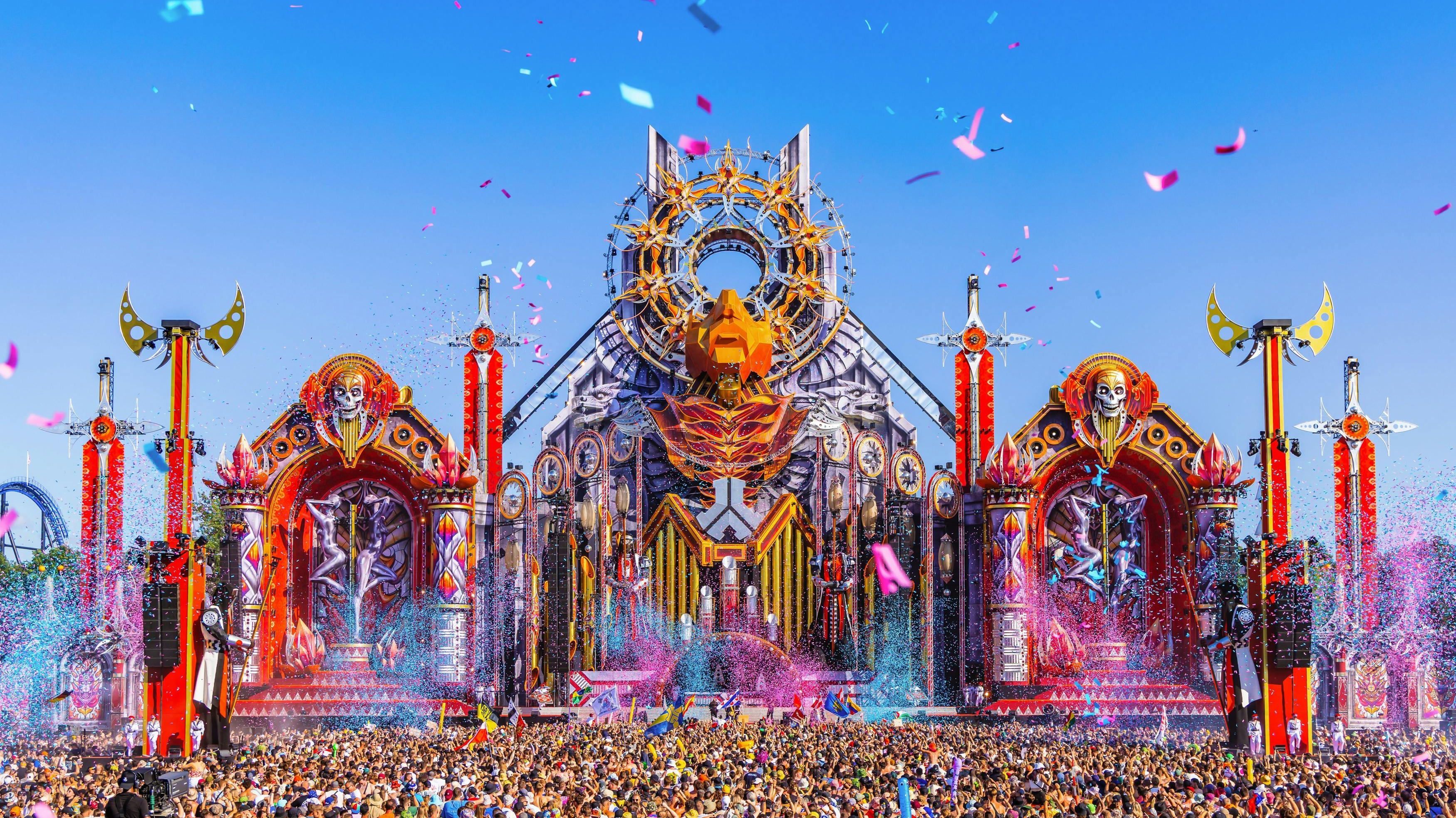 Defqon.1 festival main stage with intricate design, confetti, and a massive crowd under a clear blue sky.