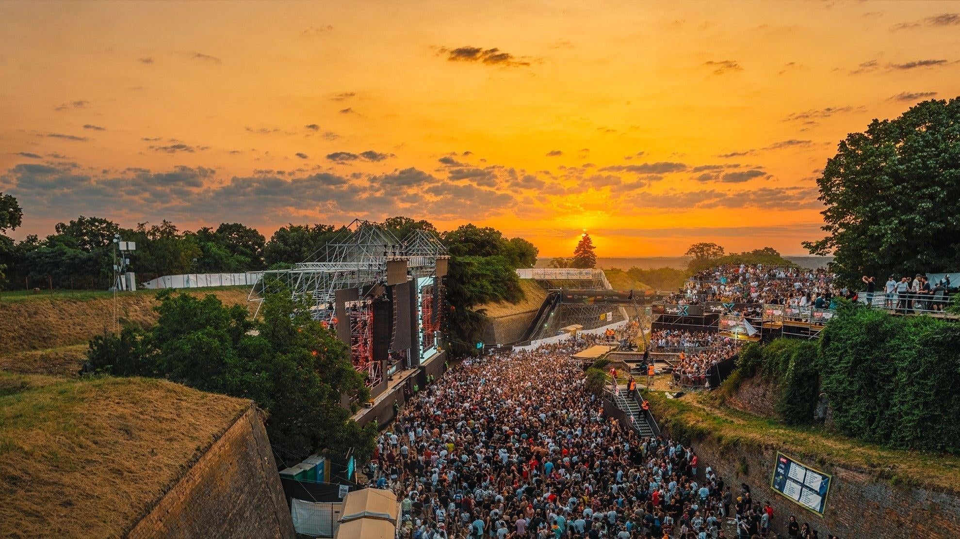 EXIT Festival crowd at sunset, main stage glowing under an orange sky.