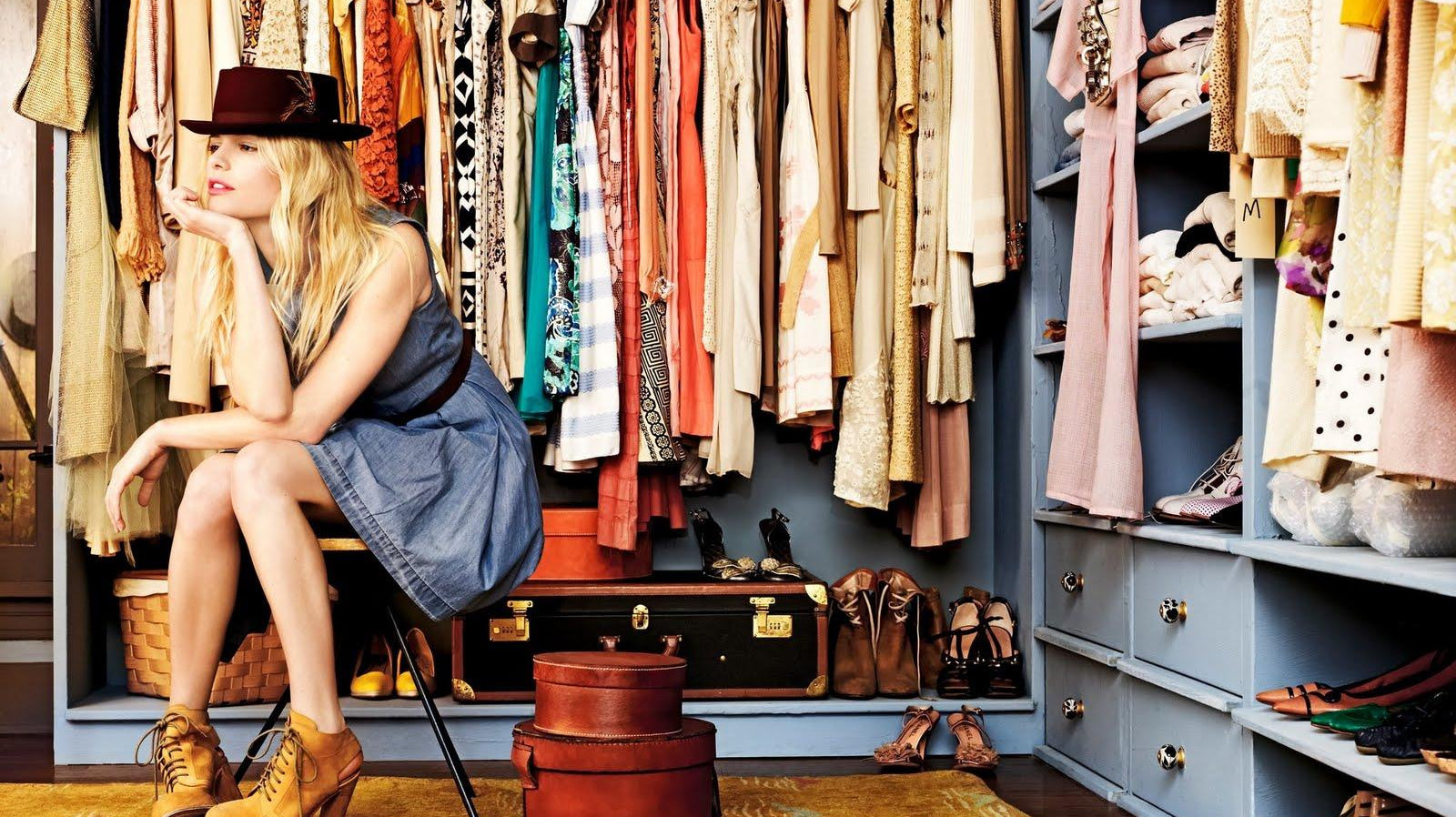 Blonde woman in a denim dress and fedora sits in a large closet filled with diverse festival clothing.