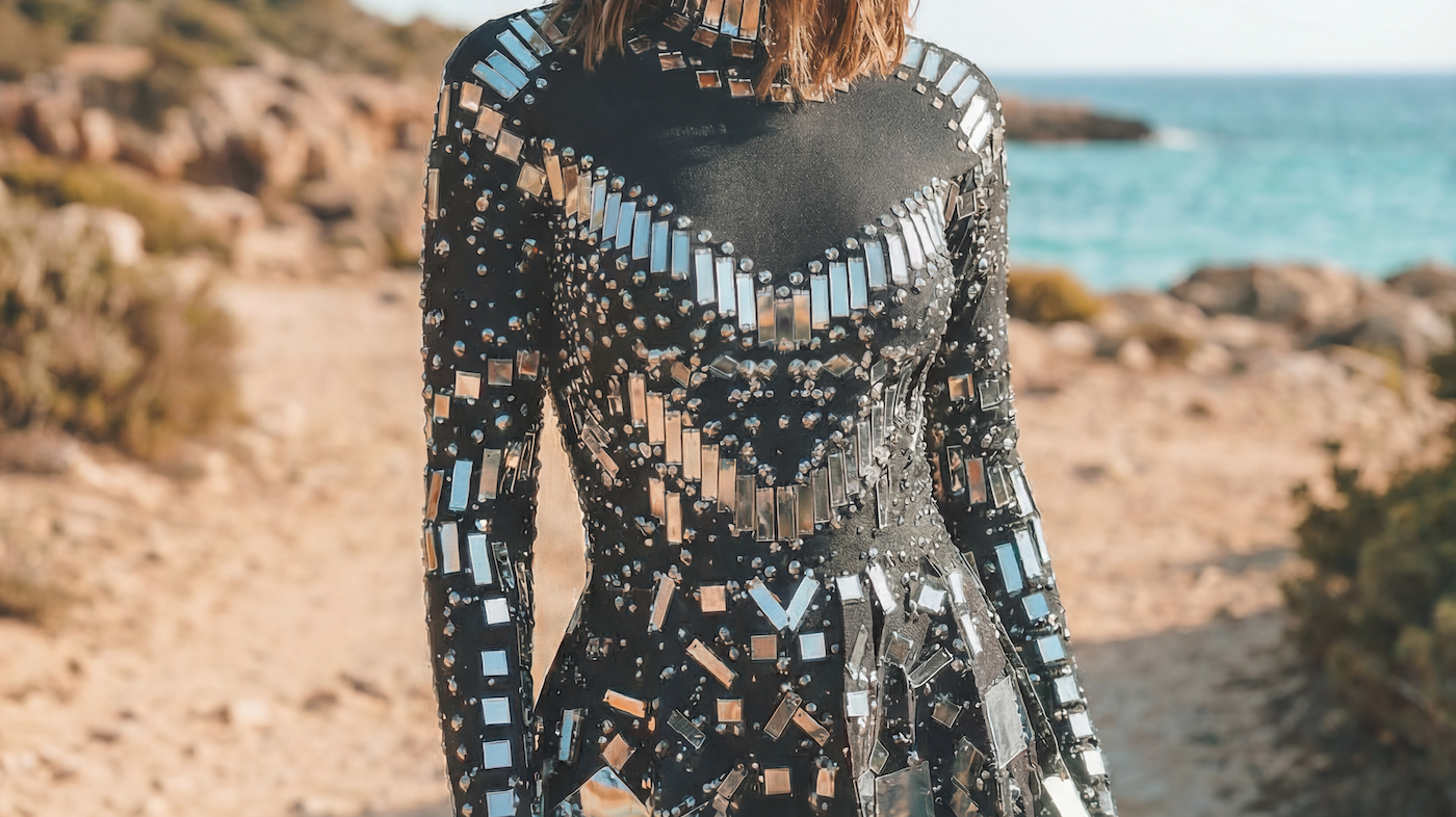 Woman in a black sequin mini dress with mirrored embellishments and a matching captain's hat on a beach path.