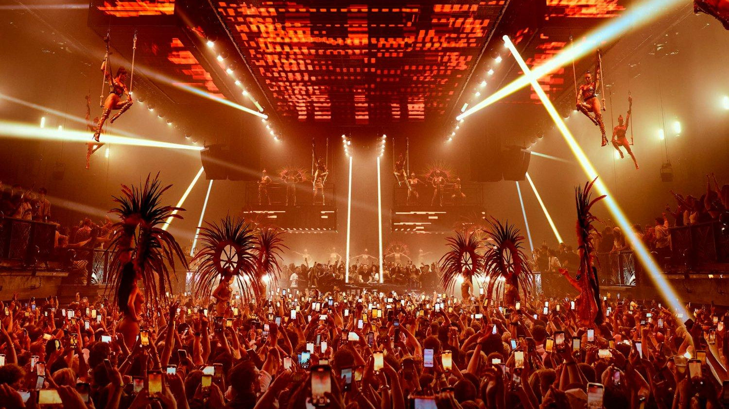 Vibrant Ibiza club scene with a large crowd, aerial performers, and red stage lighting.