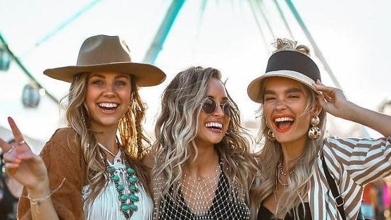 Three smiling women in stylish festival outfits, including hats and layered jewelry, with a Ferris wheel in the background.