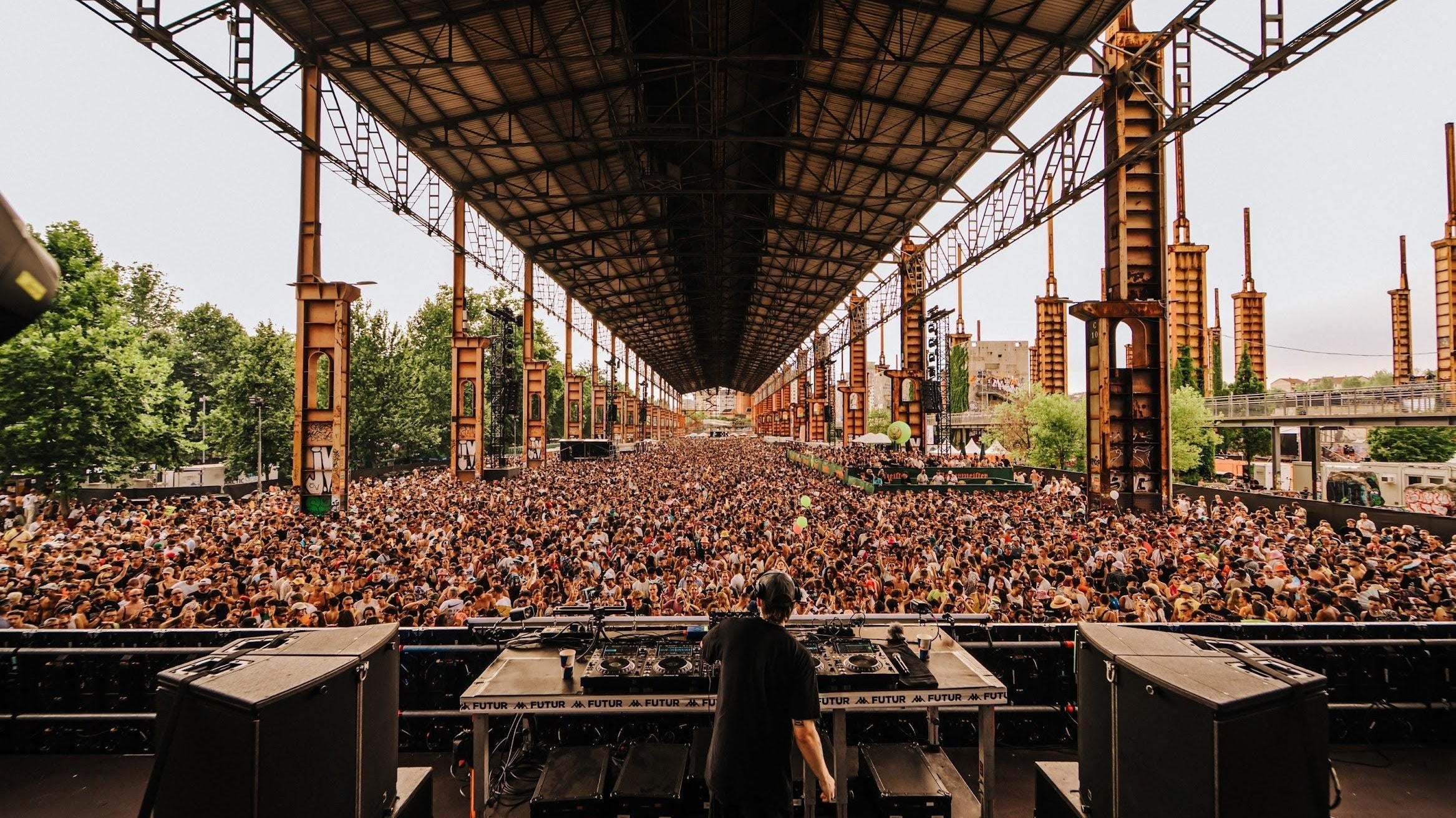 DJ on stage facing a massive crowd under a long industrial structure at Kappa FuturFestival.