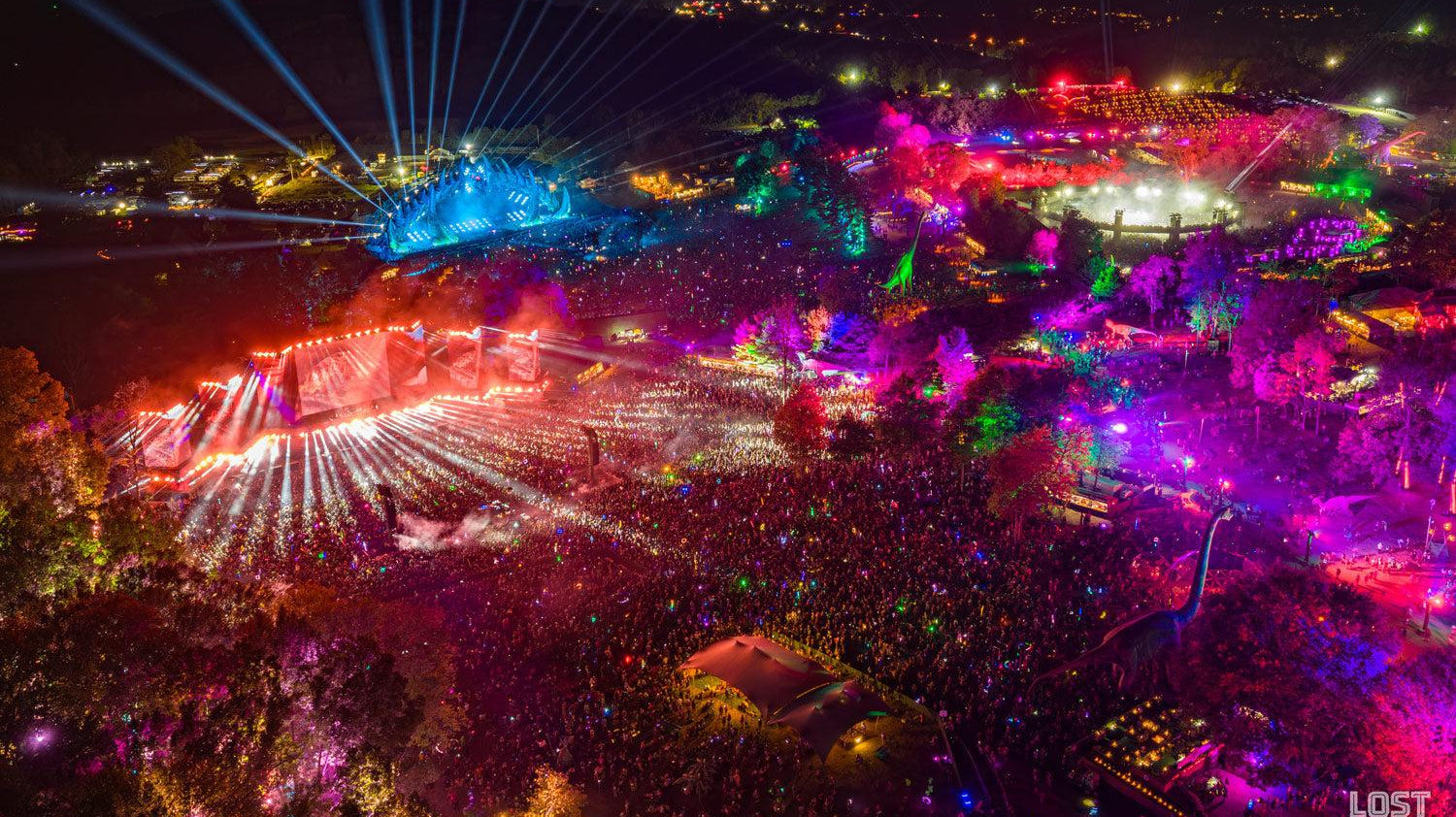 Aerial view of a massive outdoor music festival at night, with vibrant laser lights illuminating large crowds and stages.