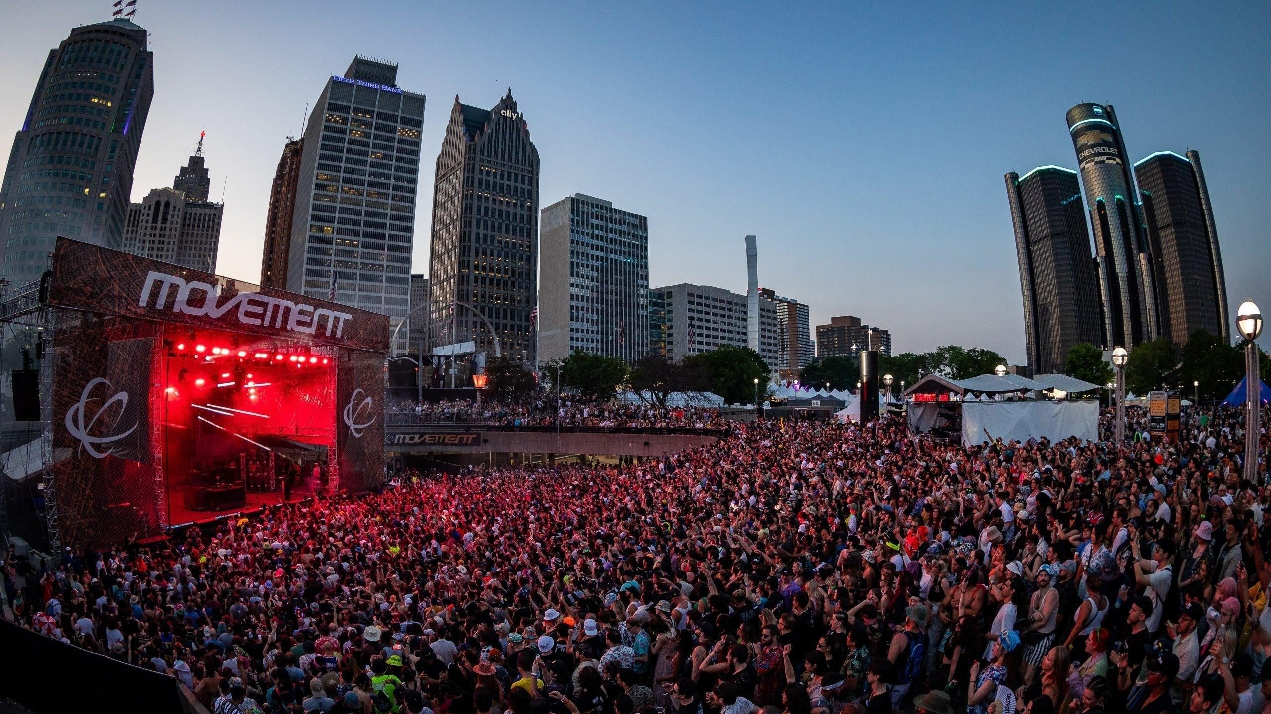 Crowd at Movement Detroit festival in Hart Plaza with city skyline at dusk, red stage lights.