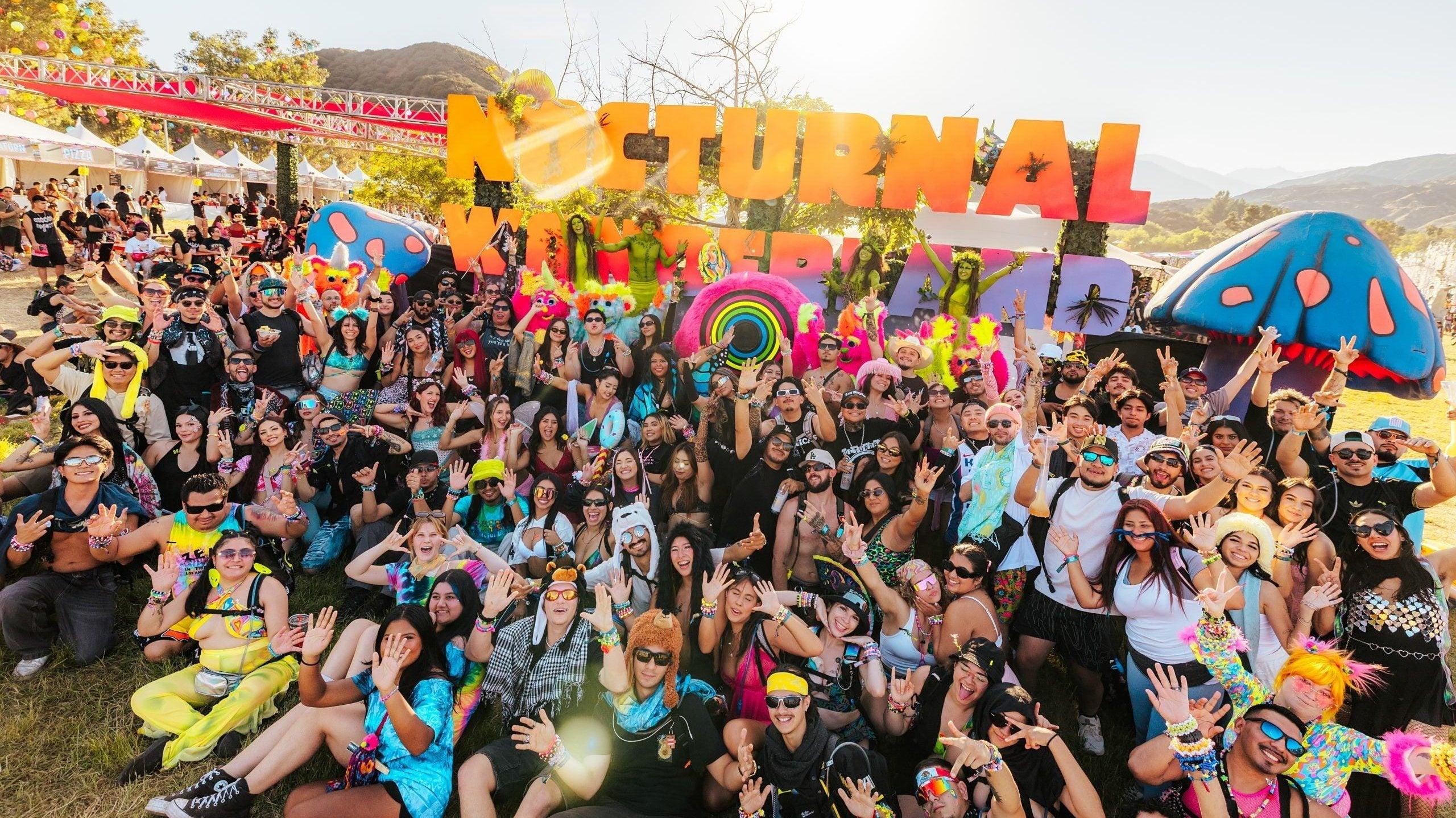 Large crowd of festival-goers posing in front of "Nocturnal Wonderland" sign and colorful mushroom art.