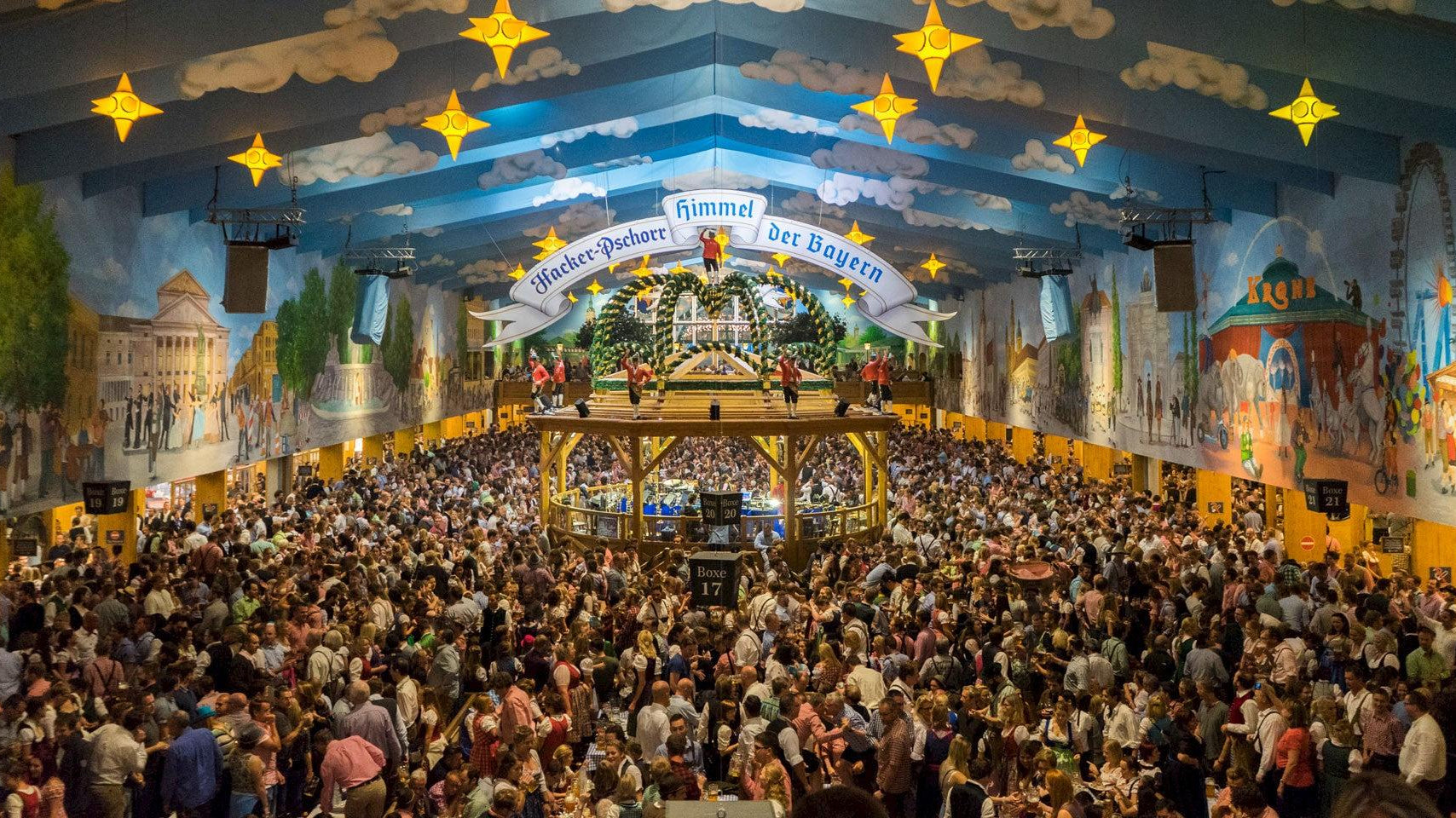 Crowded Oktoberfest beer tent with a blue sky ceiling, yellow stars, and Bavarian murals.