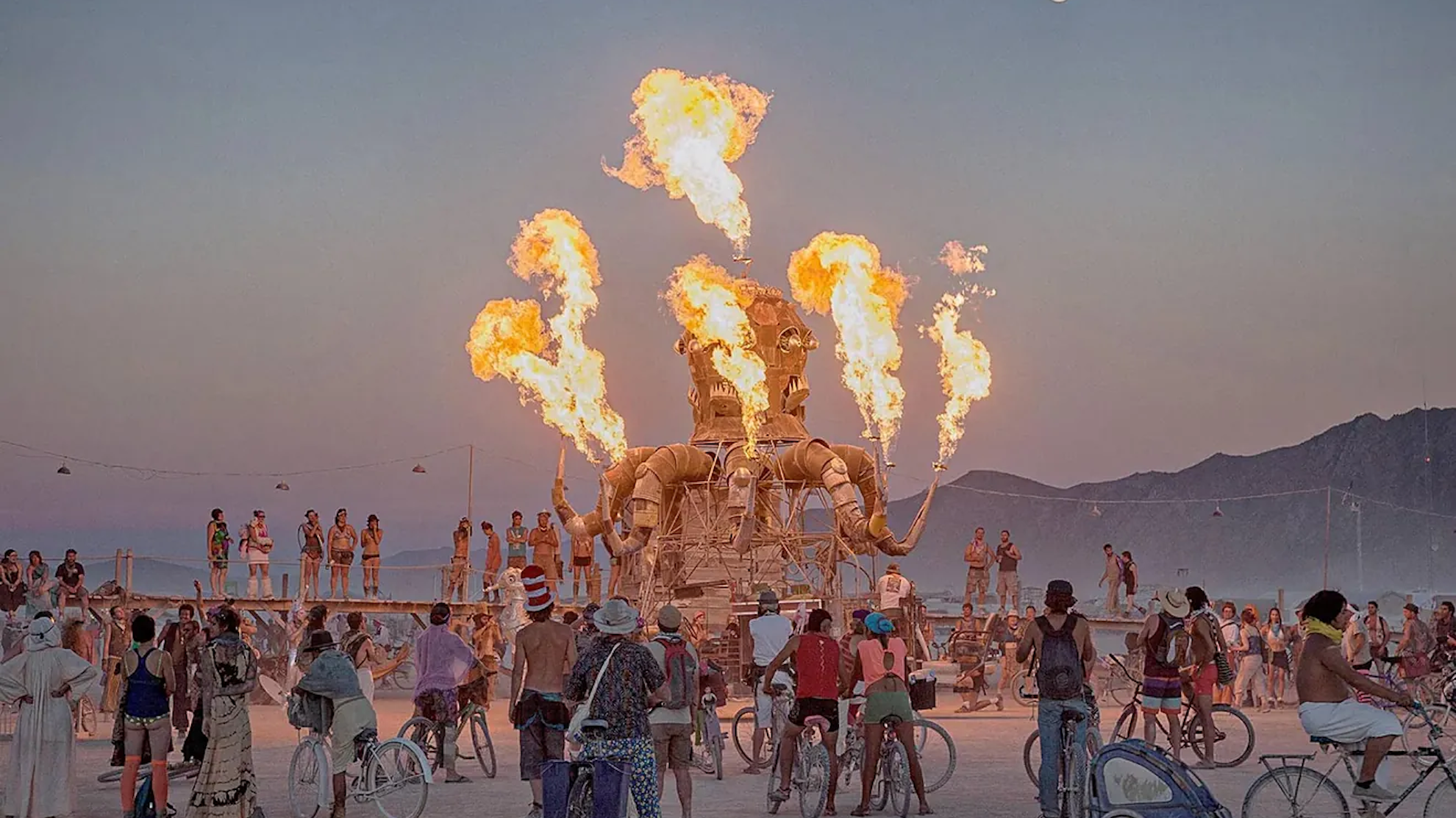 Festival-goers gather around a large, burning octopus sculpture under a full moon at dusk.
