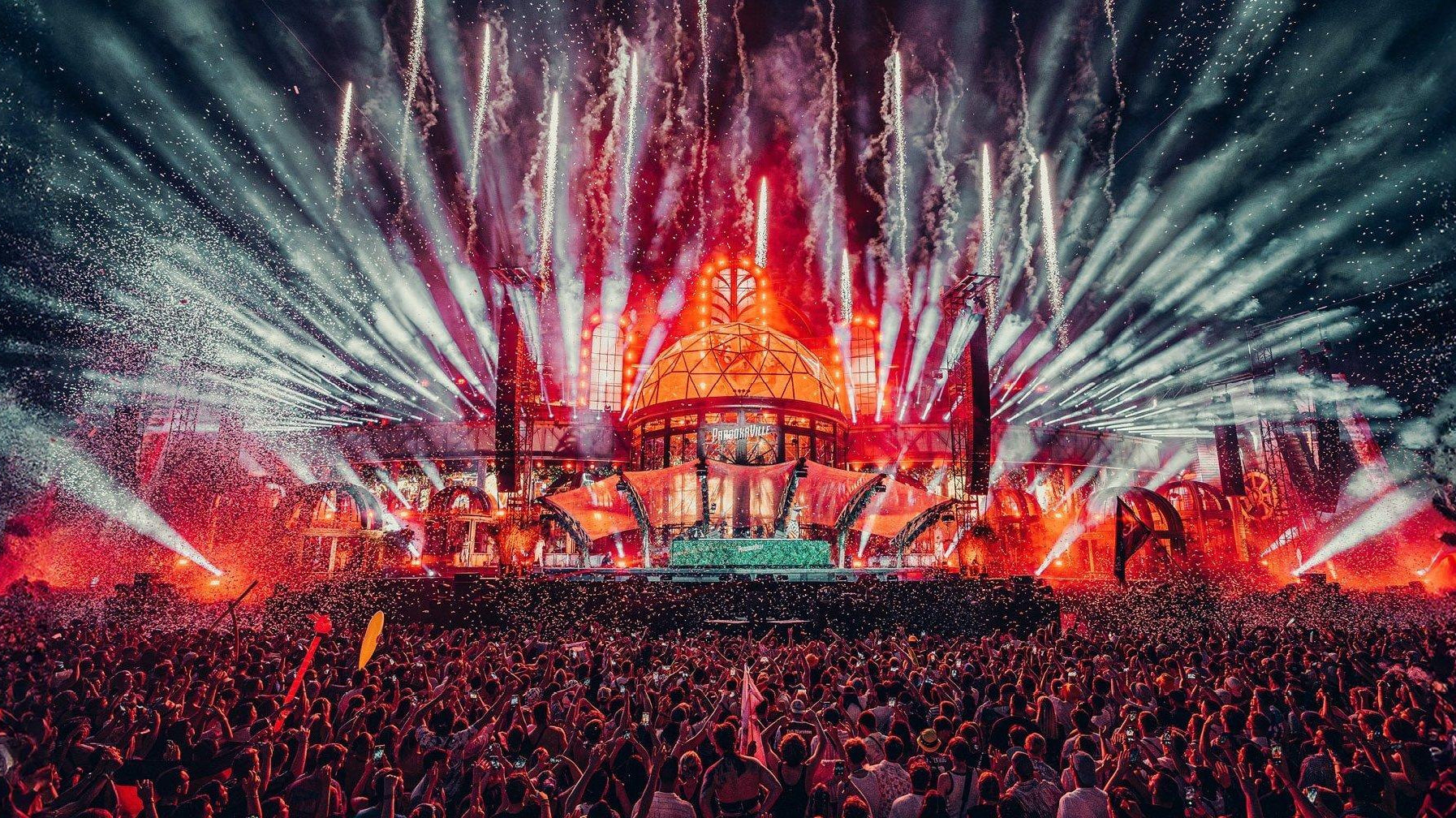 Massive outdoor festival stage with a large dome, vibrant red and white fireworks, and a huge crowd.