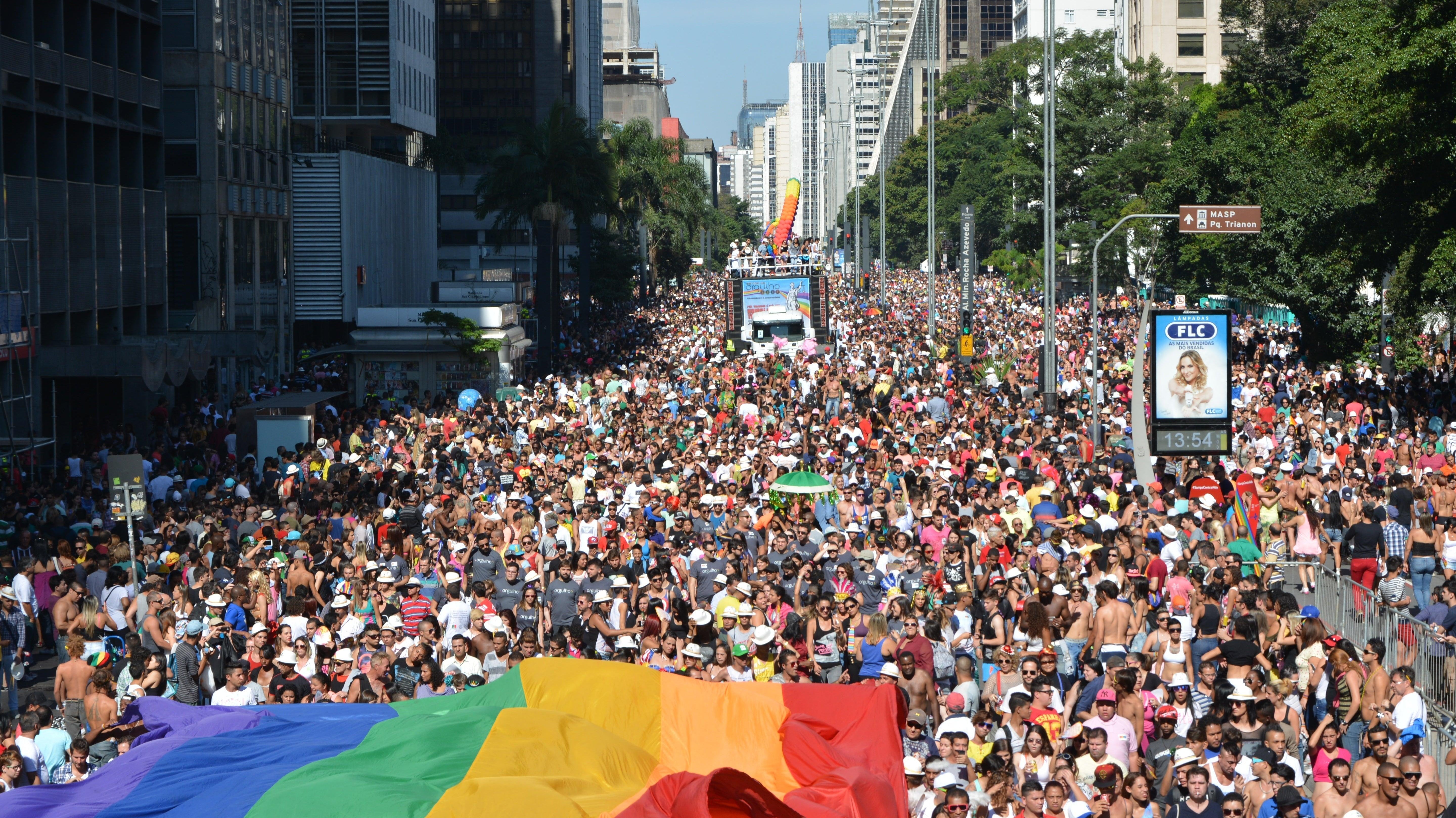 Massive crowd at São Paulo Pride, with a large rainbow flag in the foreground and parade floats.