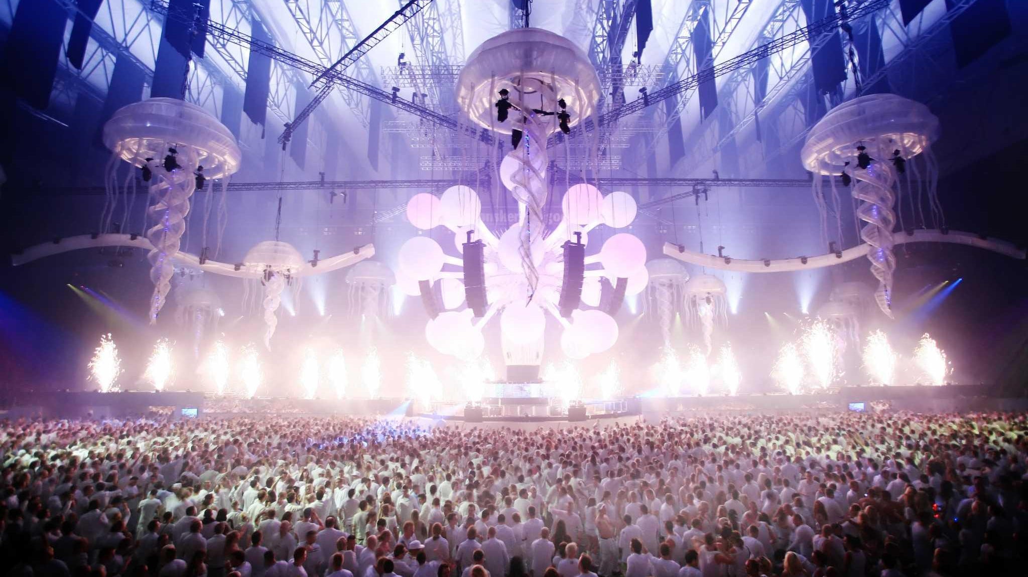 Vast crowd in white attire at Sensation Amsterdam, under glowing jellyfish decor and bright stage lights.
