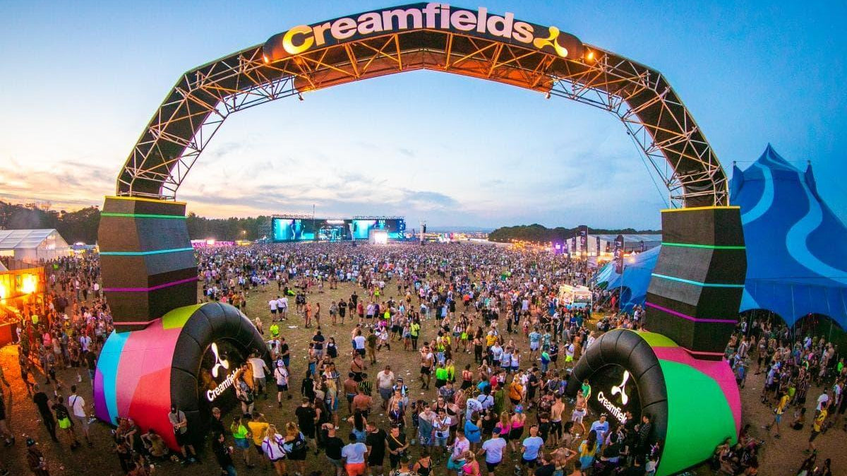 Creamfields festival entrance arch shaped like headphones, glowing at dusk over a large crowd.