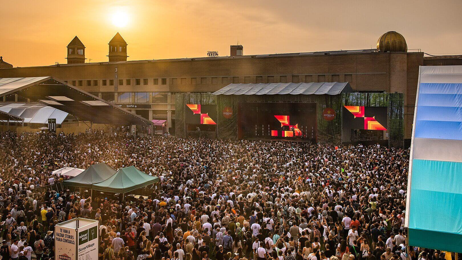 Crowd at an outdoor electronic music festival with a large stage and sunset glow.