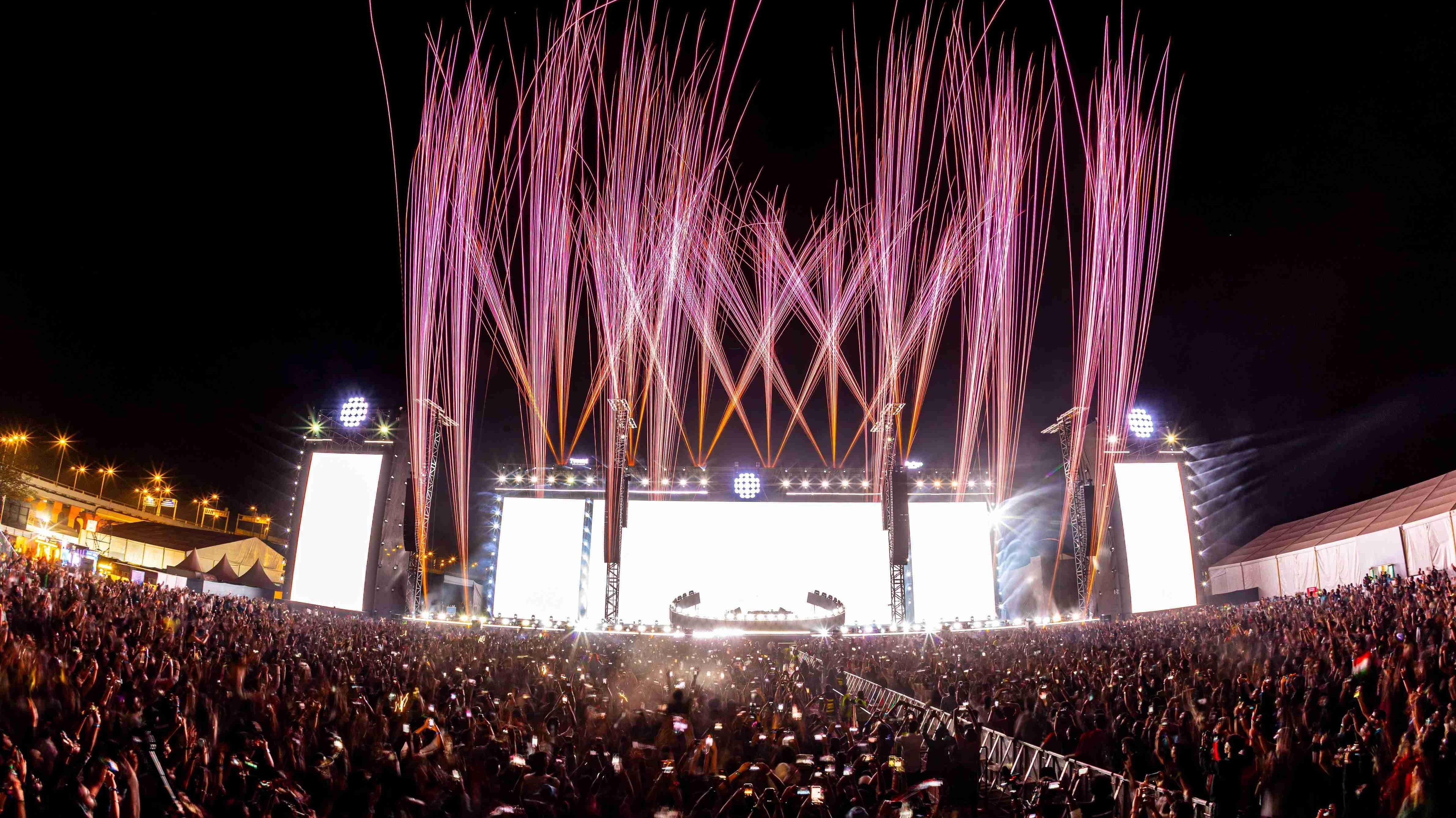 Massive outdoor EDM festival stage with bright screens, pink fireworks, and a huge crowd at night.