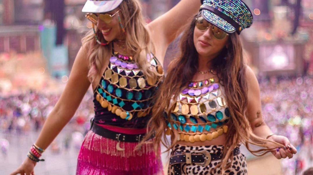 Two women in vibrant festival outfits: one in a pink fringe skirt, the other in leopard print shorts.