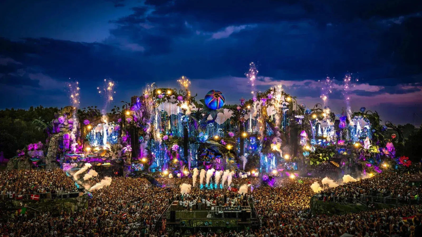 Tomorrowland main stage at dusk, vibrant with purple and blue lights, fireworks, and a massive crowd.