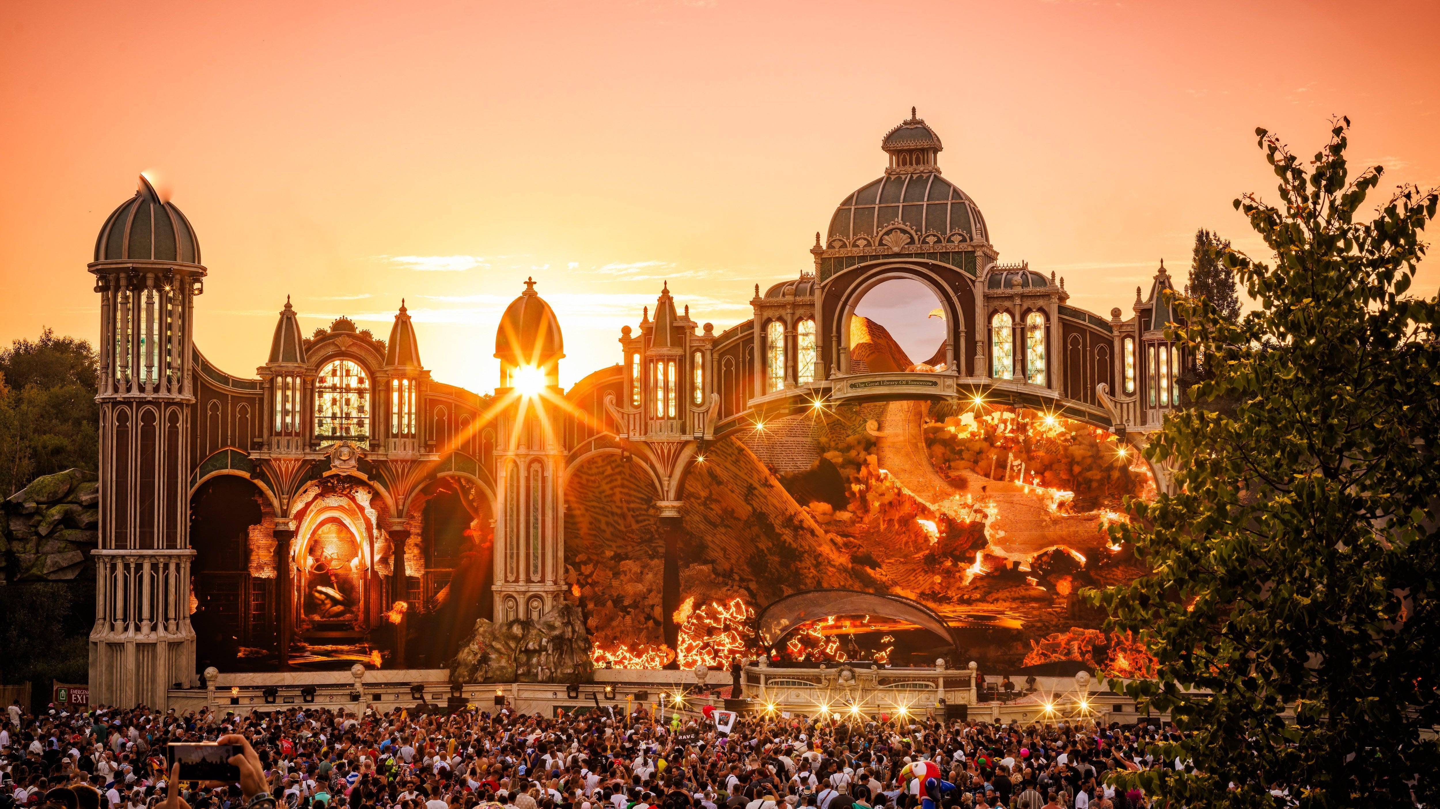 Tomorrowland main stage at sunset, with a large crowd and vibrant orange sky.