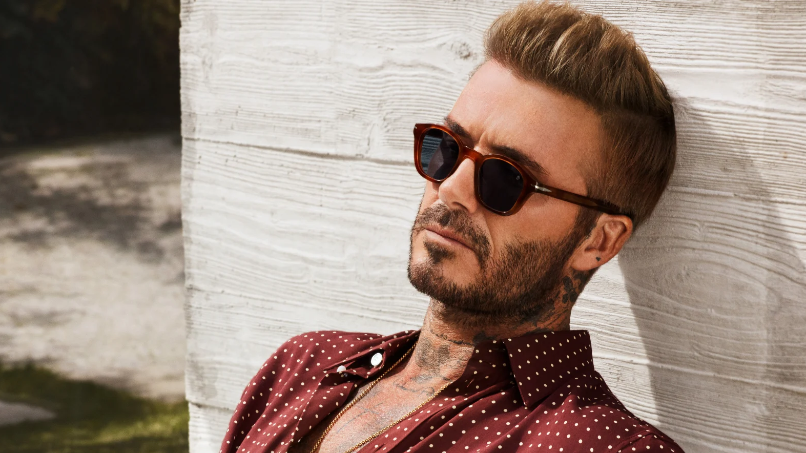 David Beckham in stylish brown tortoiseshell sunglasses, a red polka dot shirt, leaning on a textured wall.