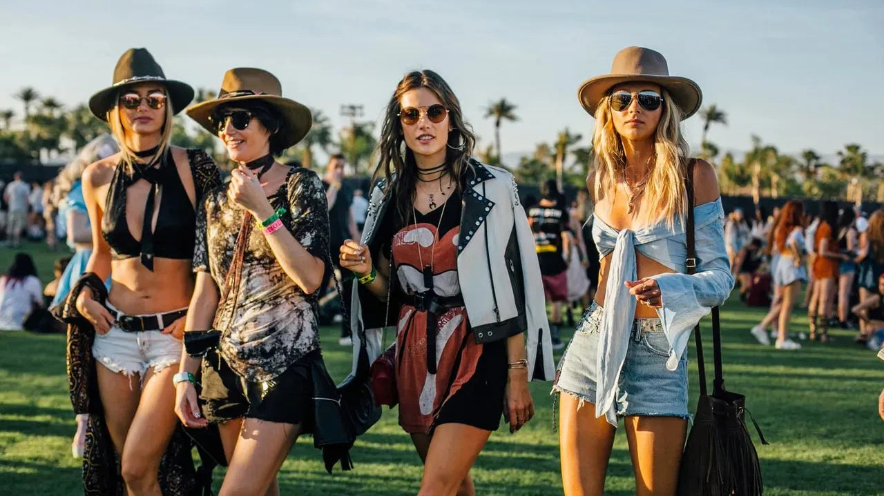 Four women in stylish festival outfits, including hats, sunglasses, and layered clothing, walking on grass.