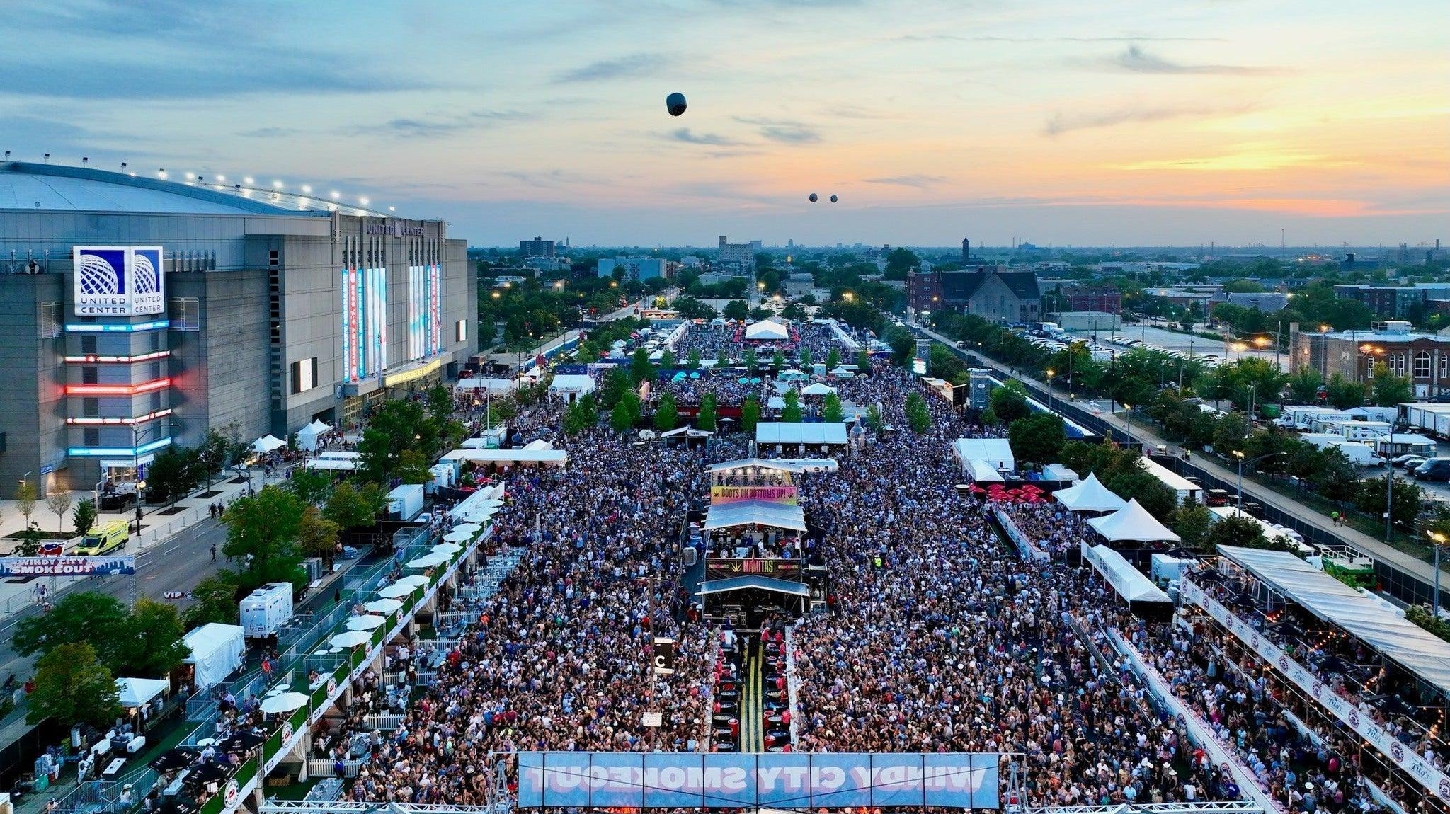 Aerial view of a massive outdoor country music festival crowd at dusk, with a stage, tents, and the United Center.
