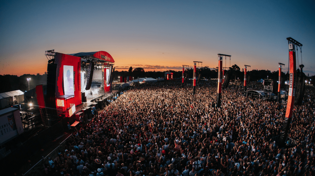 Vast crowd at an outdoor music festival with a large red stage and sunset sky.
