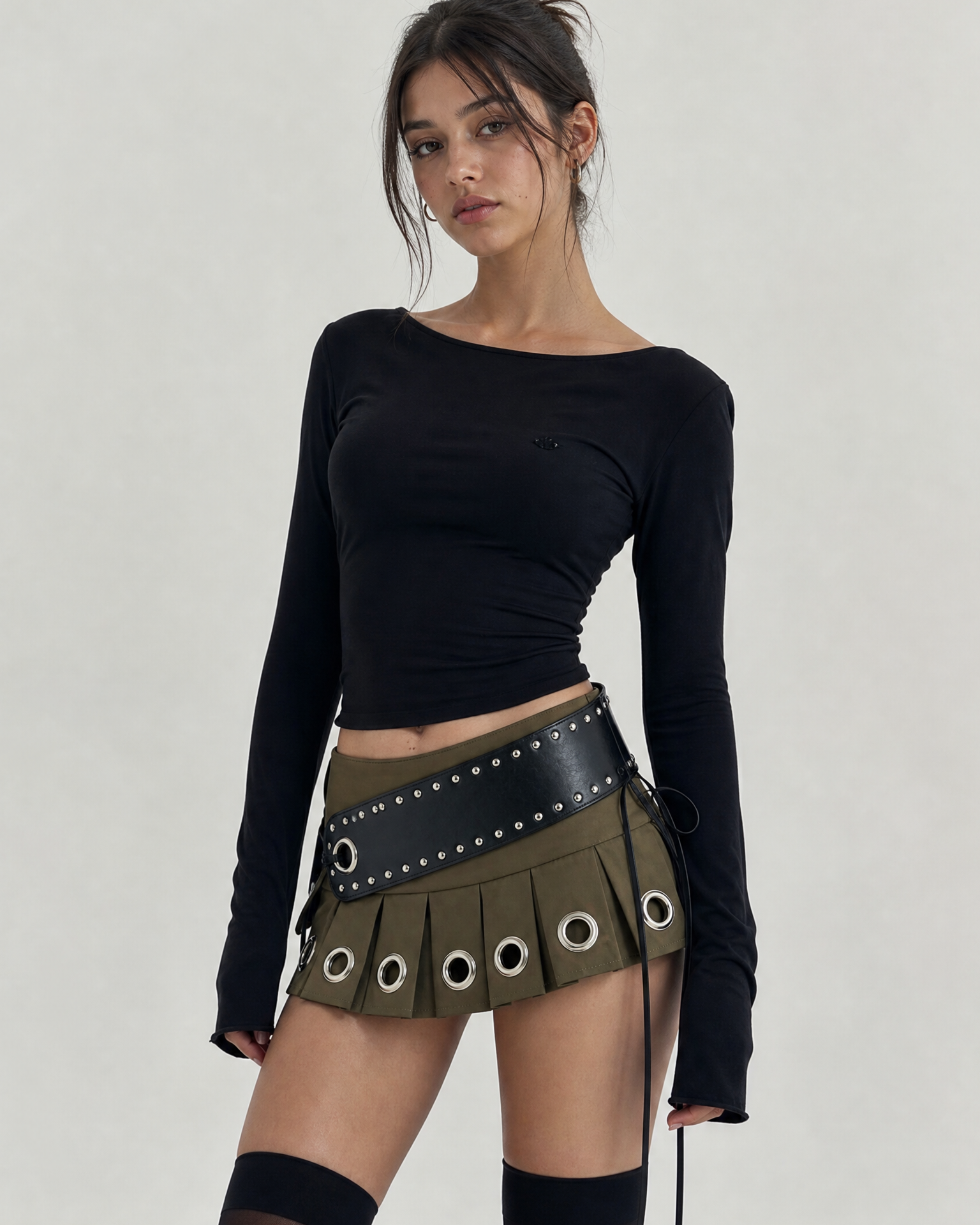 Model in a black long-sleeve top and an olive green mini skirt with large silver grommets and a studded black belt.