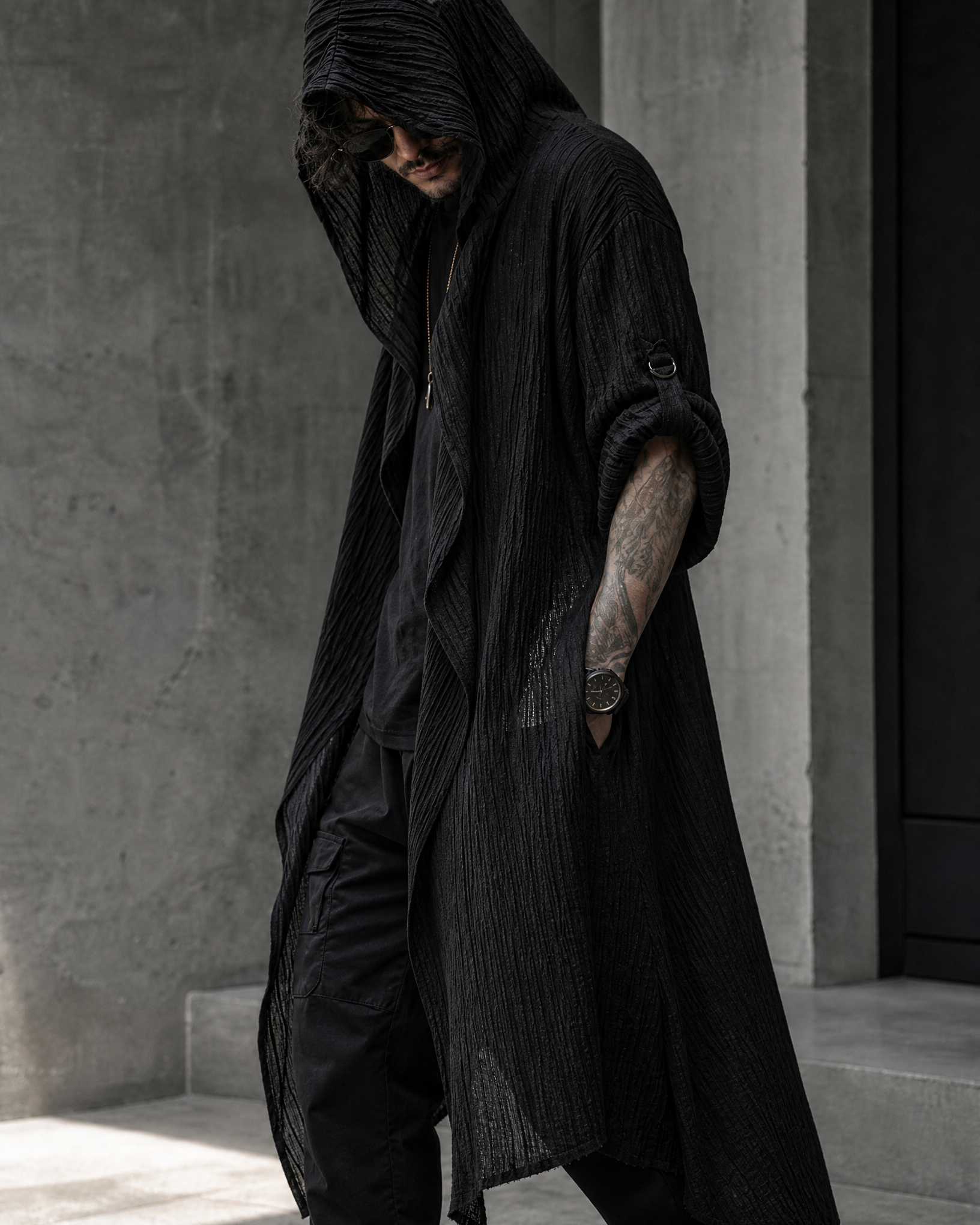 Man wearing a long, black hooded polyester cardigan with a textured finish and rolled sleeves in a modern urban setting.