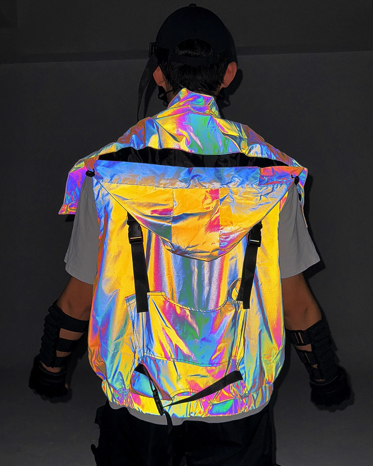 Multicolor reflective techno vest with a large hood and black strap details, worn by a person from behind.