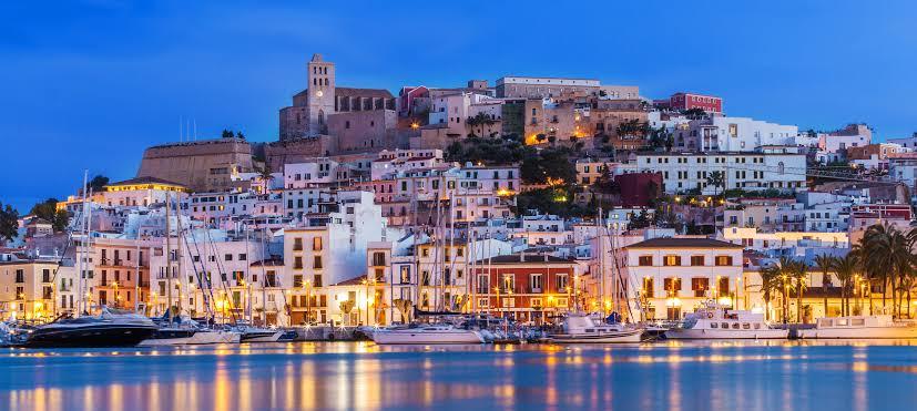 Dalt Vila's historic white buildings and cathedral illuminated at dusk above the Ibiza harbor with moored yachts.