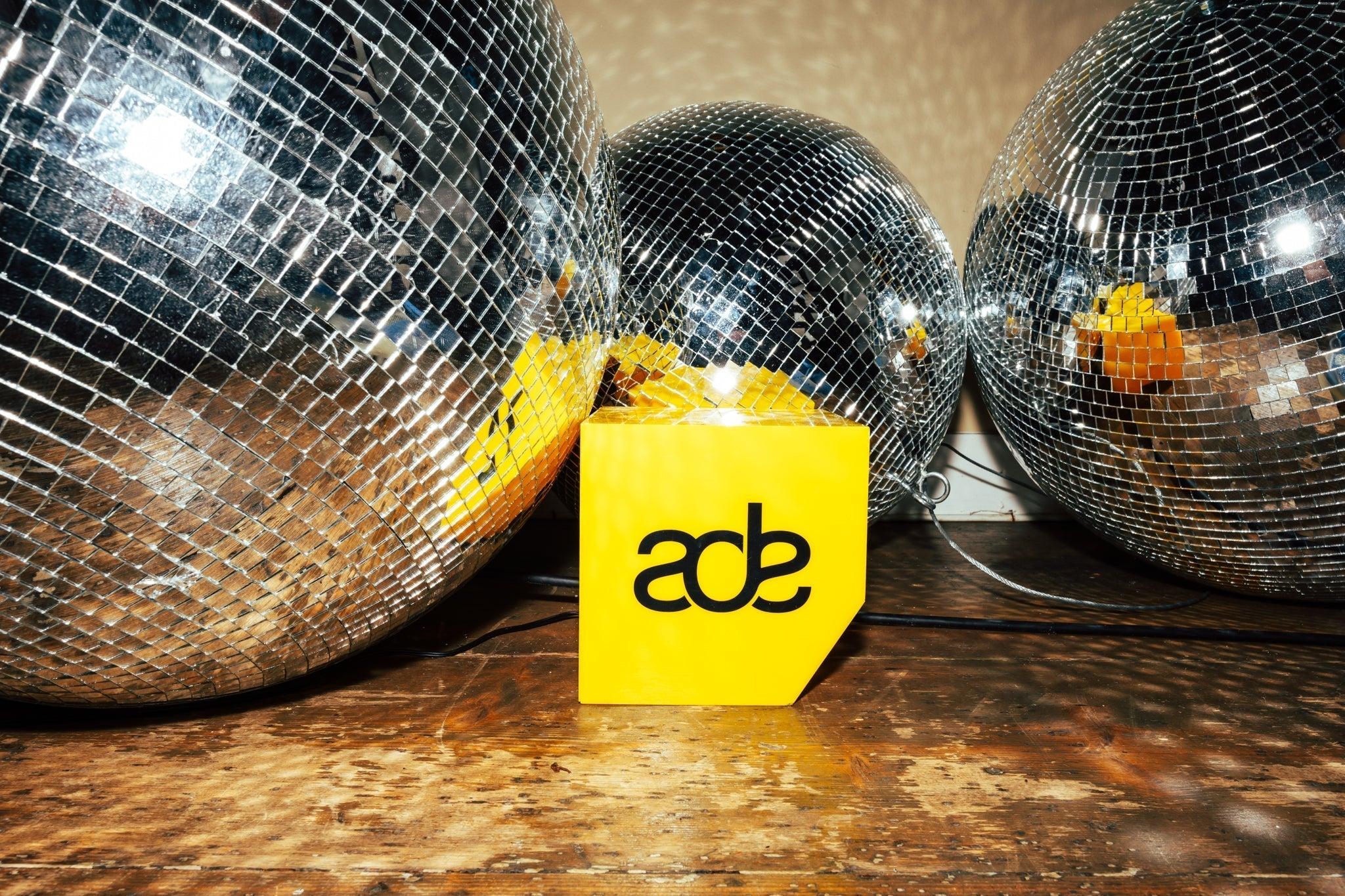 Three large disco balls surrounding a yellow ADE logo block on a wooden floor.