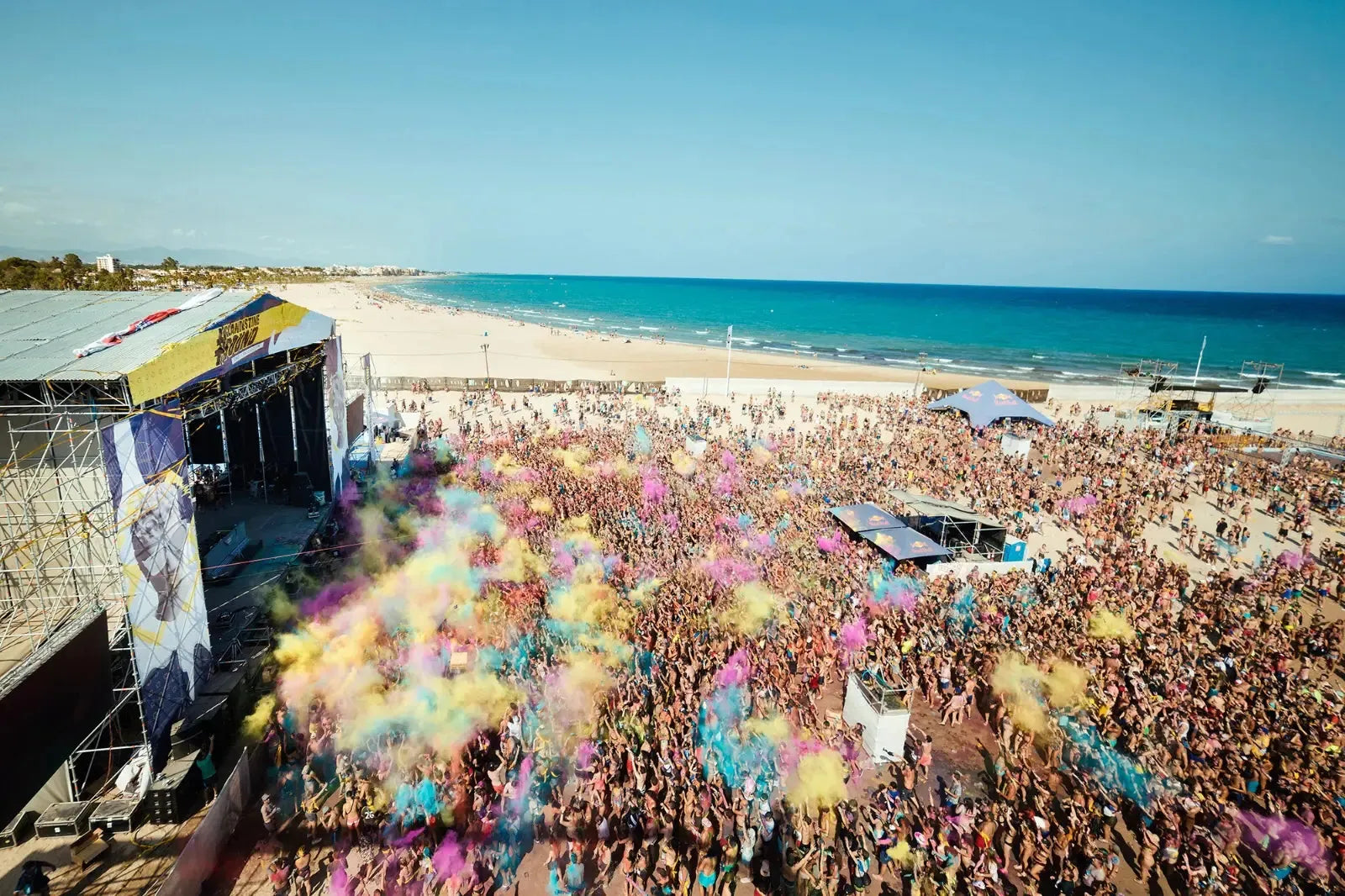 Arenal Sound beach festival with a large crowd throwing colorful powder, a stage, and the blue sea.