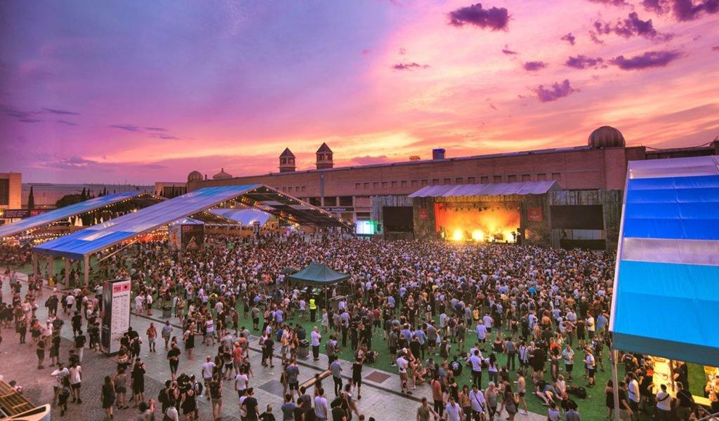 Large music festival crowd under a vibrant purple and orange sunset sky with a stage and tents.