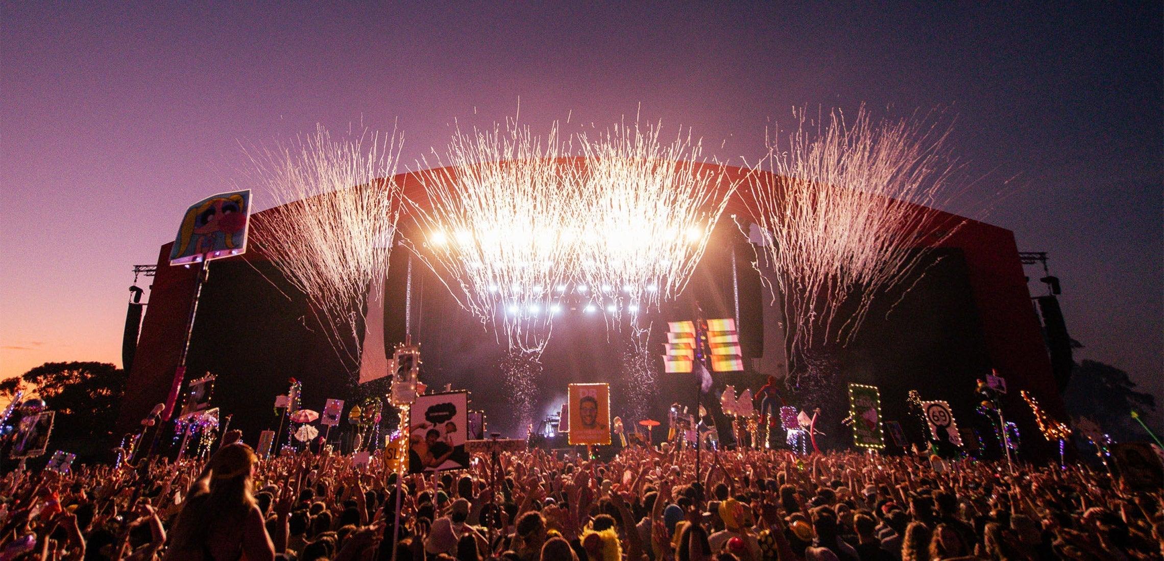 Festival crowd at sunset with fireworks bursting over a large stage, vibrant and energetic.