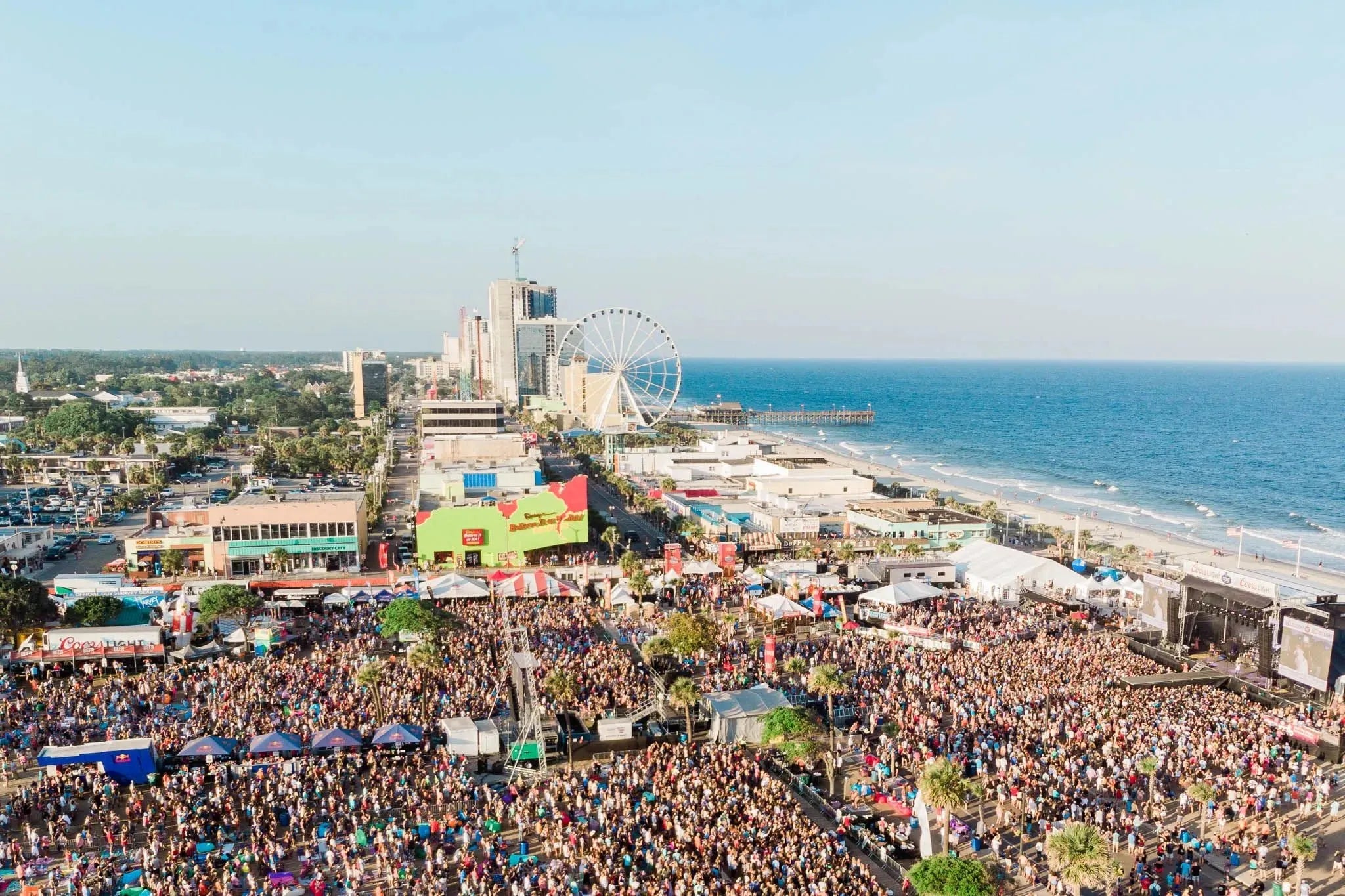 Aerial view of a crowded outdoor music festival on Myrtle Beach with a Ferris wheel and ocean.