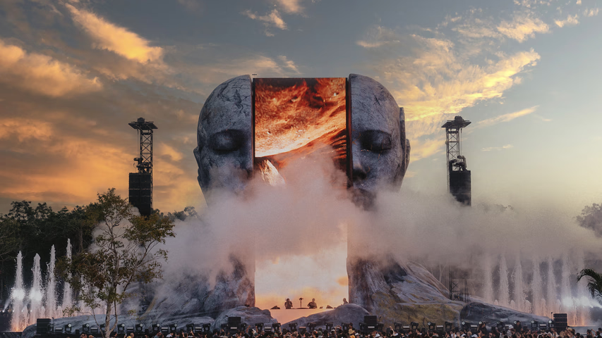 CORE Festival stage with two large stone heads, a fiery screen, mist, and water features at sunset.