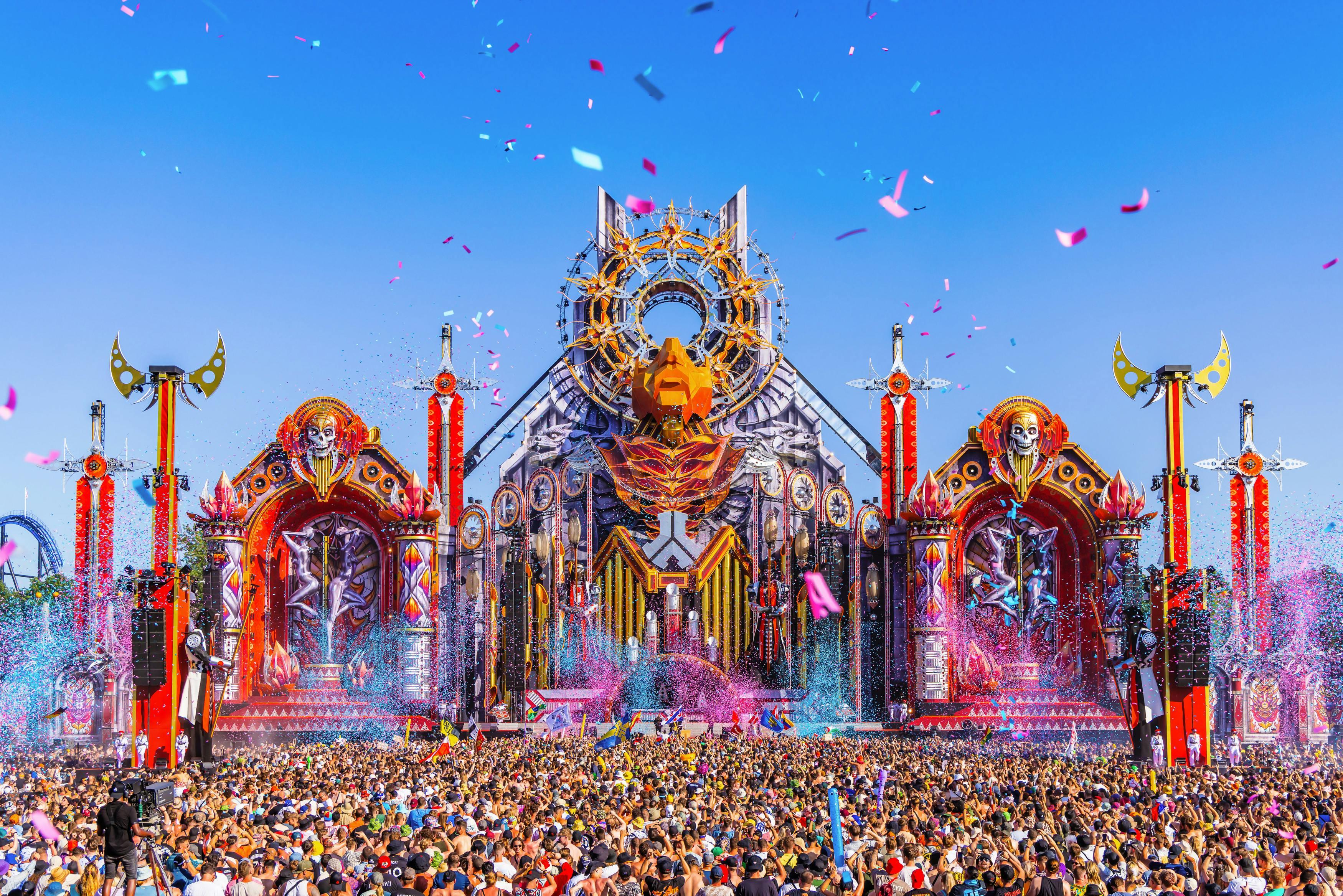Defqon.1 festival main stage with intricate design, confetti, and a massive crowd under a clear blue sky.