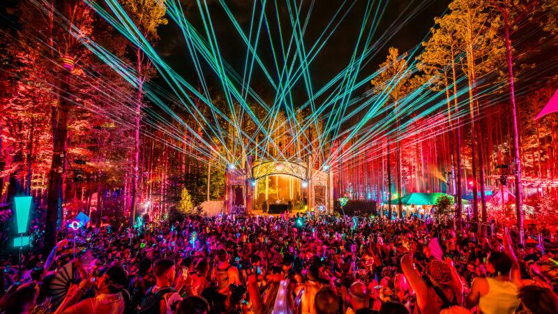 Electric Forest festival at night with vibrant green lasers over a large crowd in a colorful, illuminated forest.