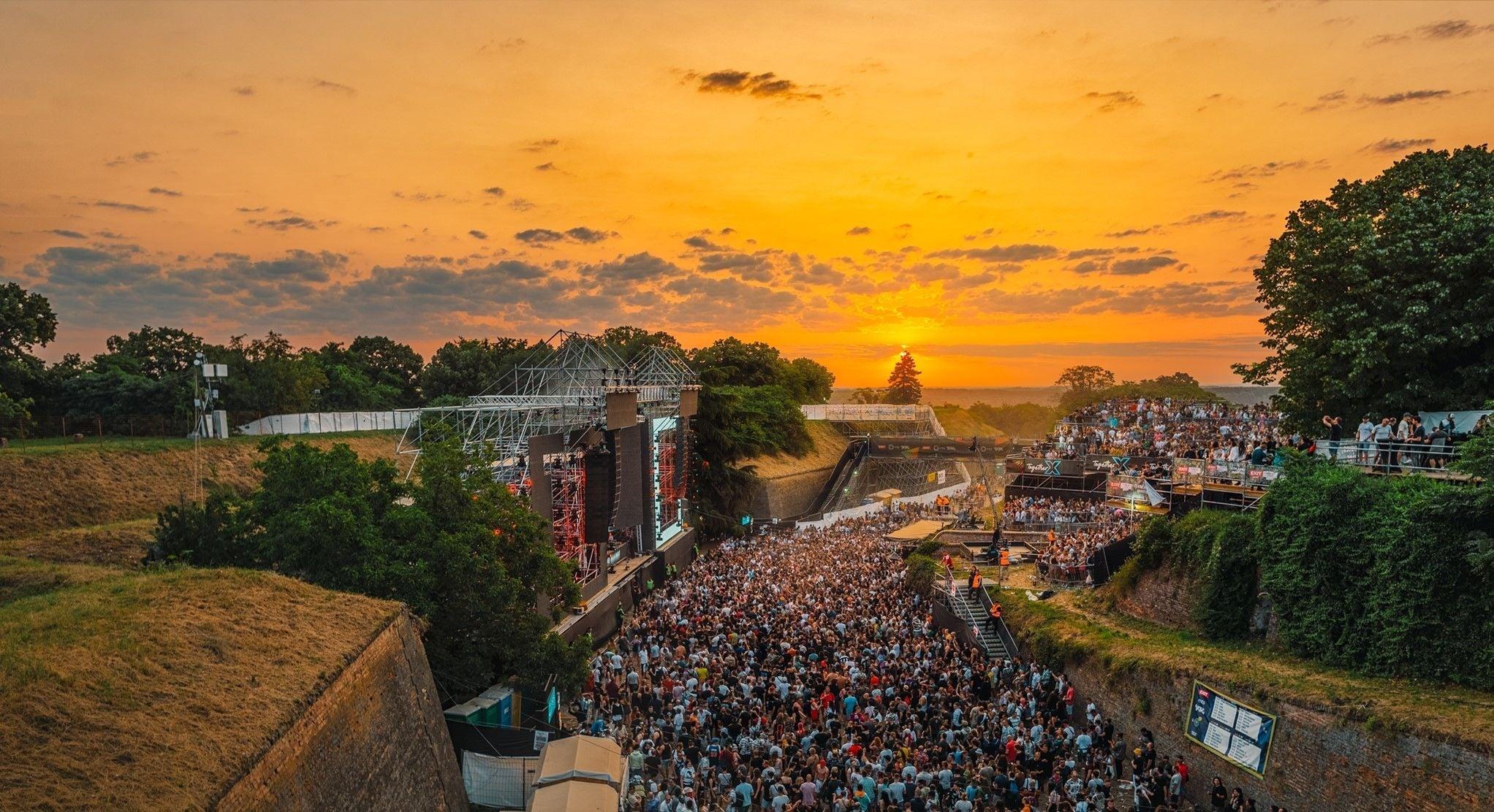 EXIT Festival crowd at sunset, main stage glowing under an orange sky.