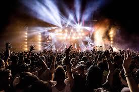 Crowd with hands raised at a vibrant outdoor music festival, illuminated by bright stage lights.