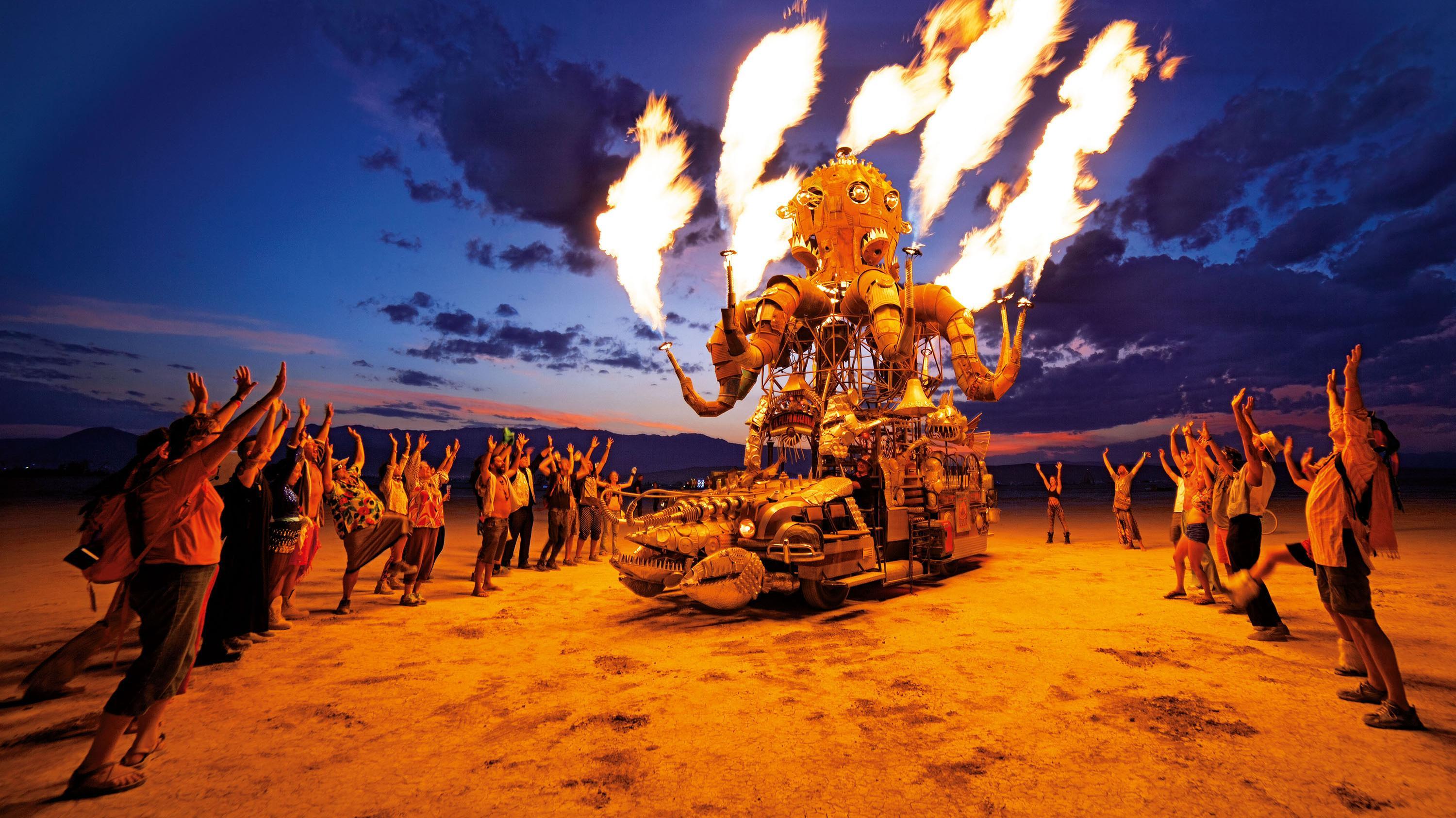 Festival-goers cheer around a flaming, steampunk-style art car at dusk, transitioning from day to night.