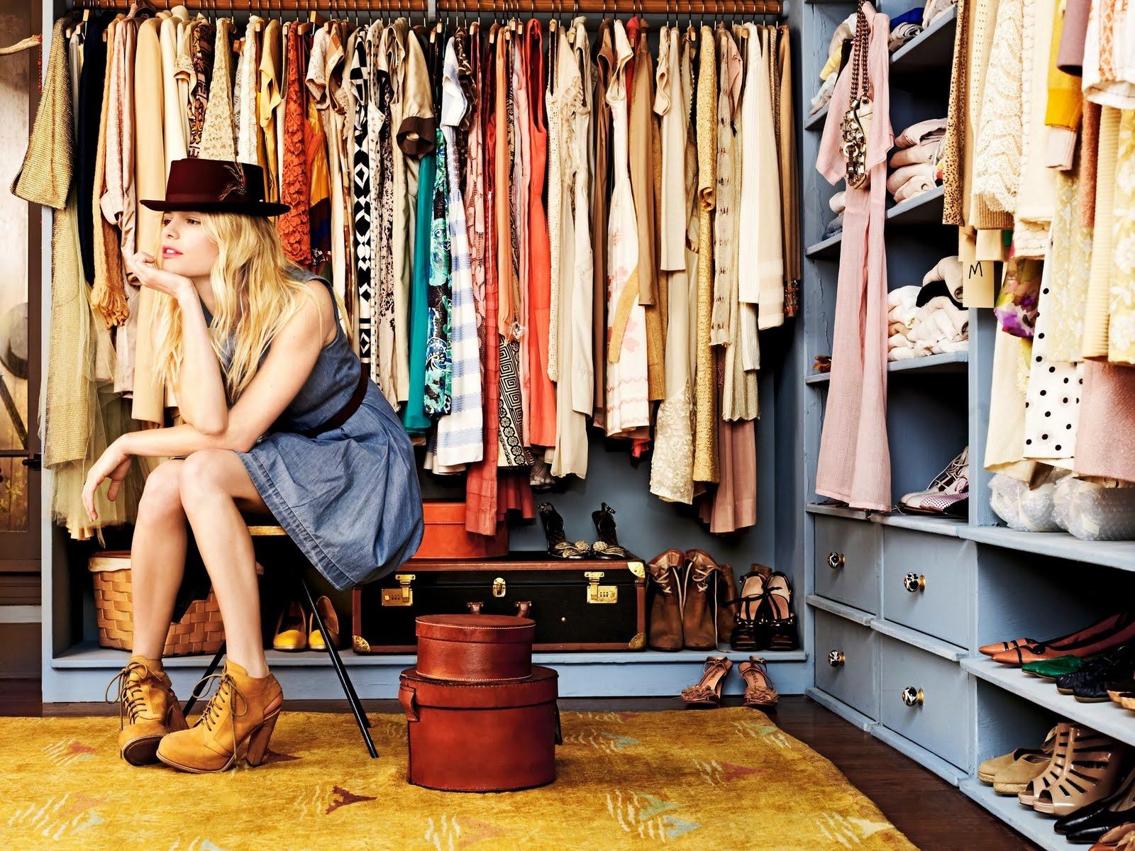 Blonde woman in a denim dress and fedora sits in a large closet filled with diverse festival clothing.