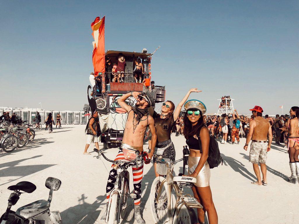 Three people in unique Burning Man outfits with bikes, a colorful art car, and dusty playa in the background.