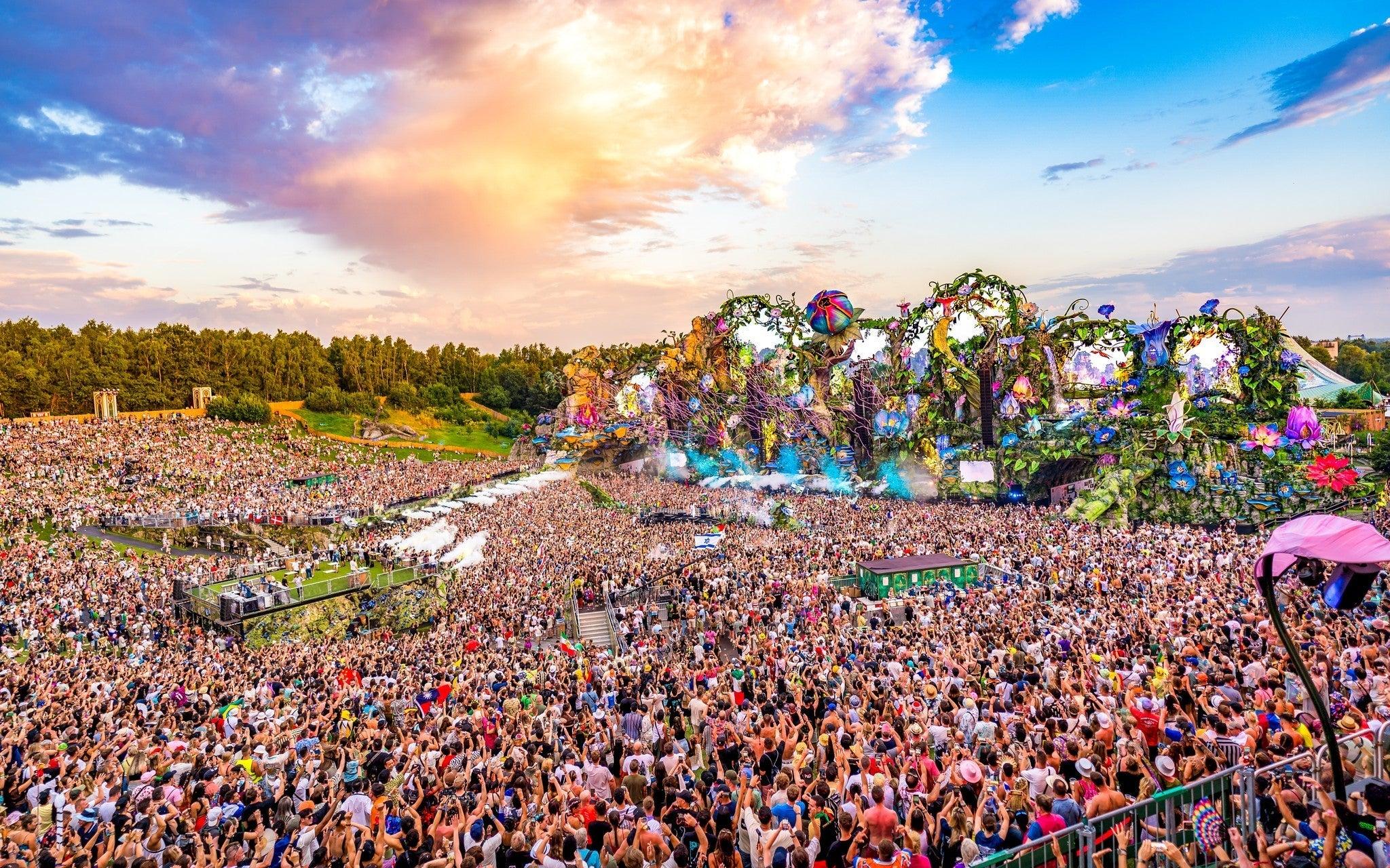 Vast festival crowd under a vibrant sunset sky, with an elaborate, colorful stage in the background.